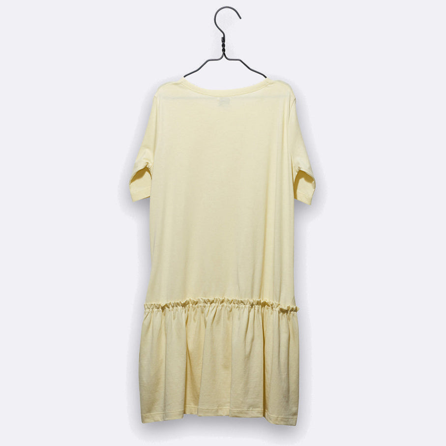 Ein hellgelbes kurzärmeliges Kleid hängt auf einem schwarzen Bügel vor einem schlichten weißen Hintergrund. Das Kleid hat eine lockere Passform und eine geraffte Rüsche am unteren Saum.