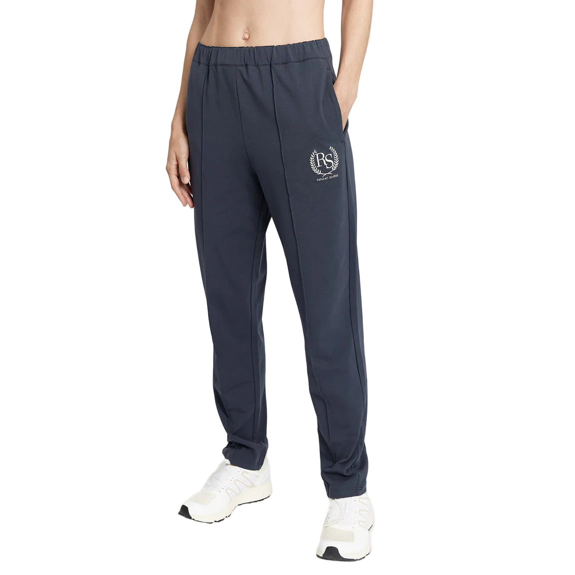 Eine Person, die eine dunkelblaue Jogginghose mit einem weißen Logo auf dem linken Oberschenkel und weiße Turnschuhe trägt, steht vor einem schlichten weißen Hintergrund.