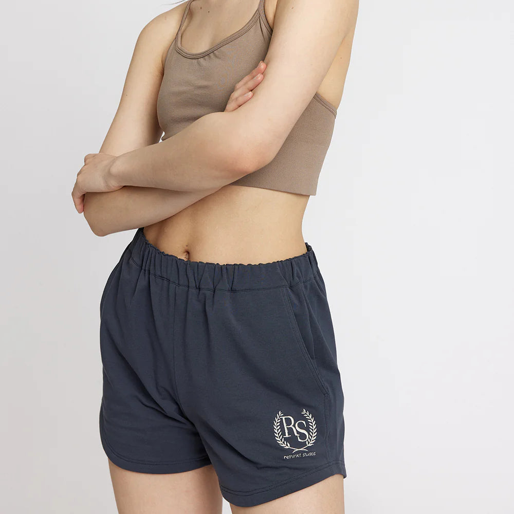 Eine junge Frau mit langen dunklen Haaren, die ein taupefarbenes Crop-Top und marineblaue Shorts mit einem weißen RS-Logo trägt, steht mit verschränkten Armen vor einem schlichten weißen Hintergrund.