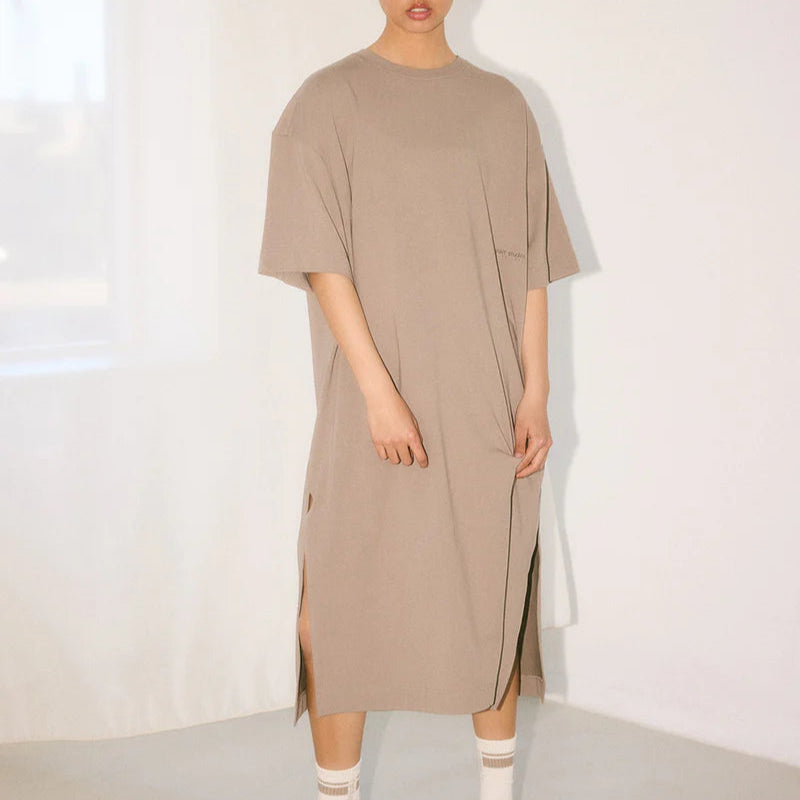Eine Person steht in einem Haus und trägt ein lockeres, hellbraunes, knielanges T-Shirt-Kleid mit Seitenschlitzen, weiße Socken und schwarze Sportschuhe. Im Hintergrund sind weiße Vorhänge und ein Fenster zu sehen.