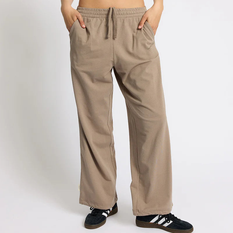 Eine Person, die ein weißes Crop-Top, eine lockere beigefarbene Hose mit Kordelzug und schwarze Turnschuhe trägt, steht mit den Händen in den Taschen vor einem schlichten hellen Hintergrund.