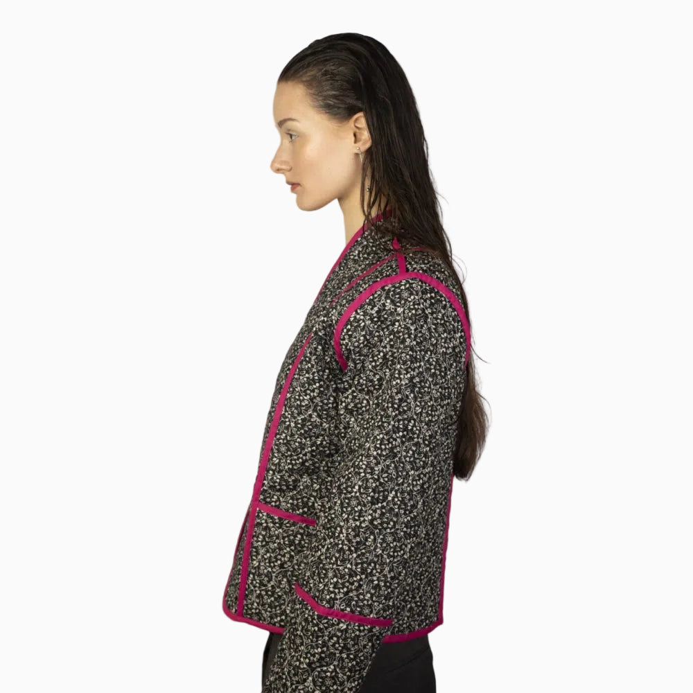 Eine Person mit langen, nassen Haaren steht im Profil vor einem unifarbenen Hintergrund und trägt eine schwarz-weiß gemusterte Jacke mit leuchtend rosafarbenem Saum.