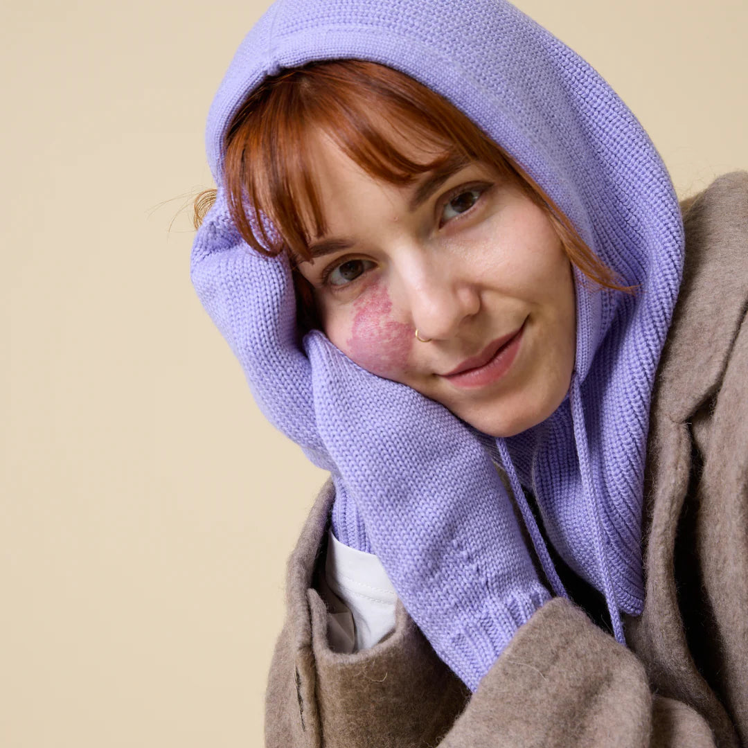 Eine Frau mit heller Haut und rotem Haar, die eine lilafarbene Strickhaube und Handschuhe trägt, stützt ihr Gesicht auf ihre behandschuhte Hand und lächelt sanft. Sie hat ein rosafarbenes Muttermal auf der linken Wange und ist mit einem hellbraunen Mantel bekleidet.