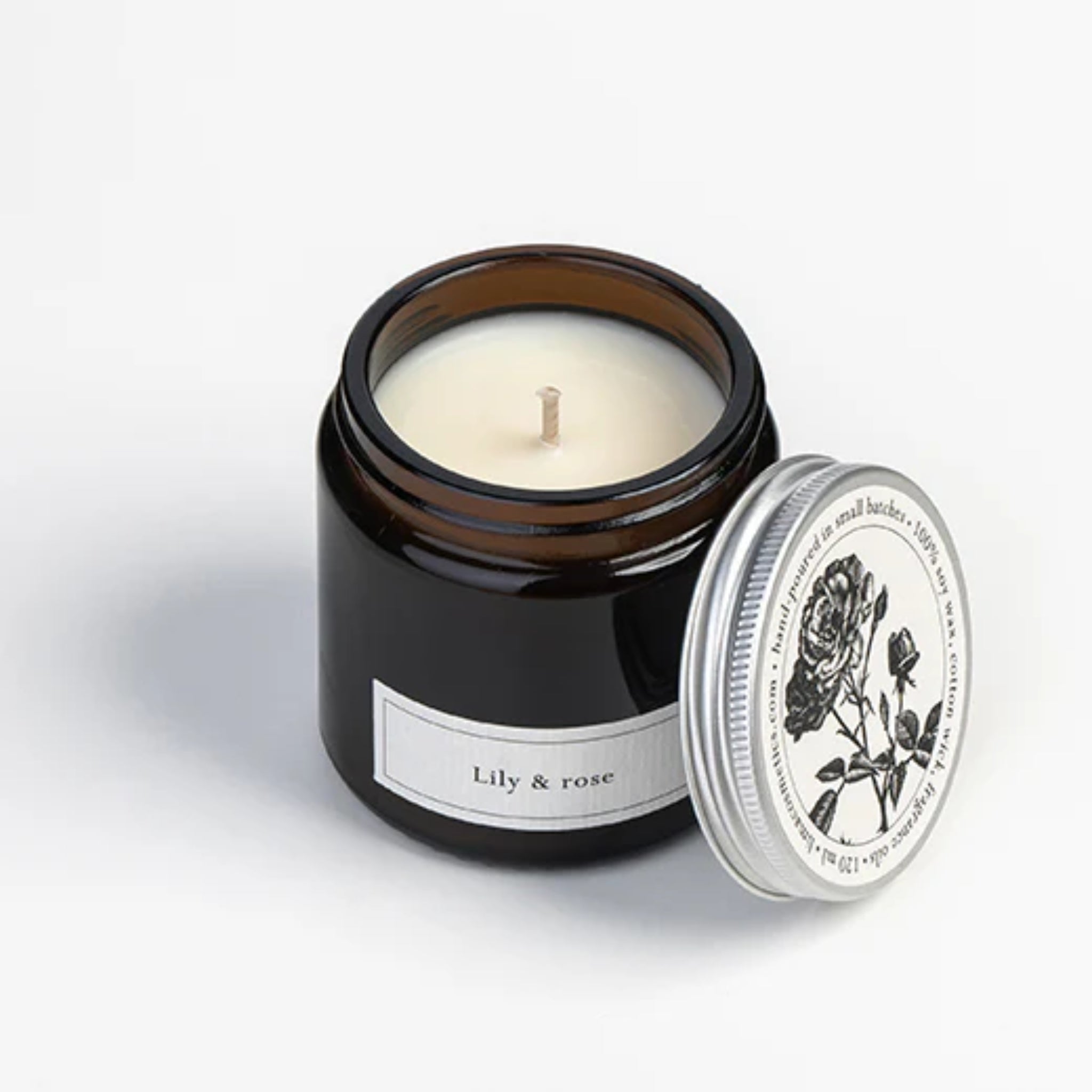 Ein braunes Glas mit der Aufschrift "Lily & Rose" und einer weißen Wachskerze im Inneren. Der silberne Deckel, der neben dem Glas ruht, zeigt eine schwarze Rosenillustration und einen dekorativen Text am Rand.