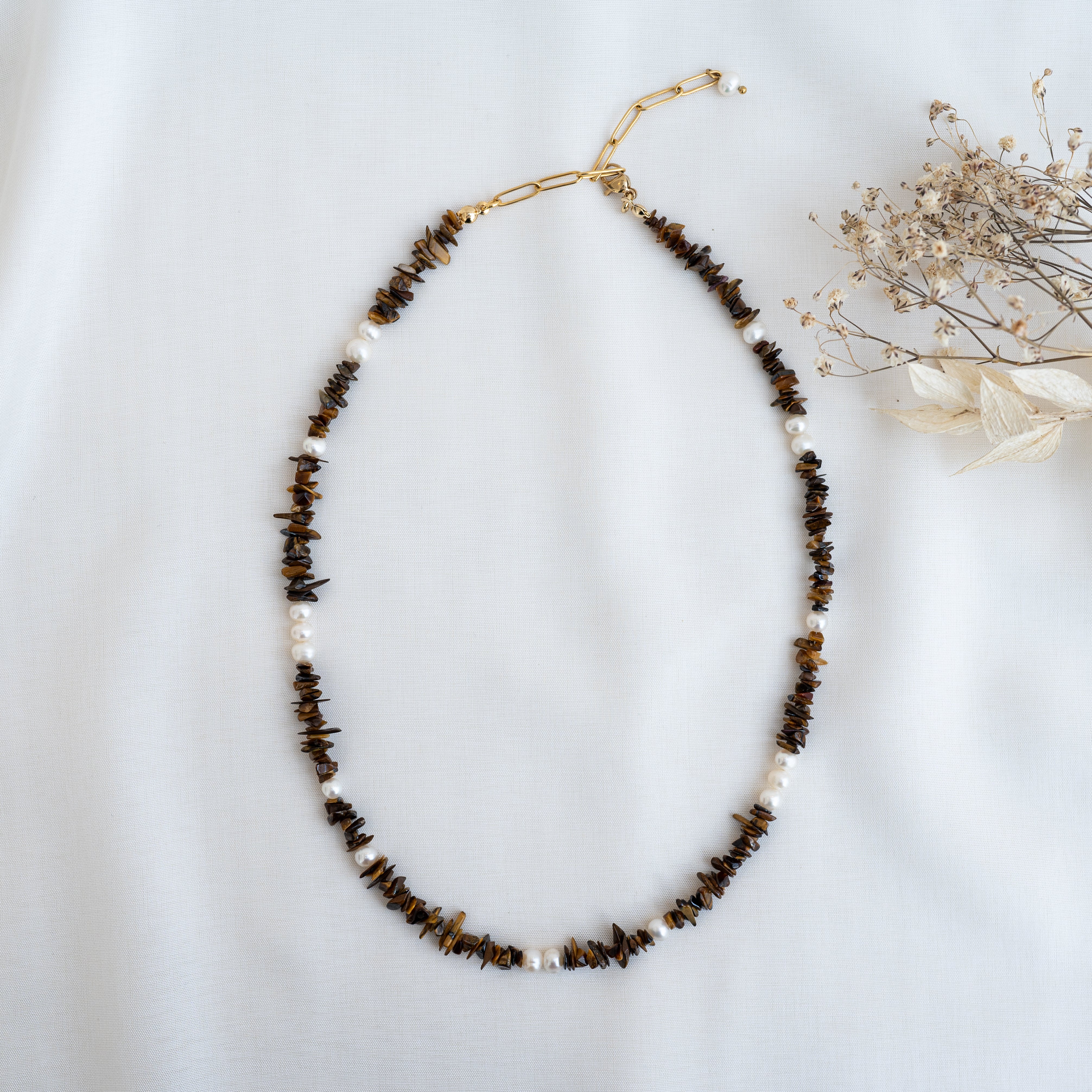 Eine Perlenkette mit braunen Steinsplittern und weißen Perlen, die mit einem goldenen Verschluss versehen ist, wird auf einem weißen Stoffhintergrund neben getrockneten Blumen präsentiert.