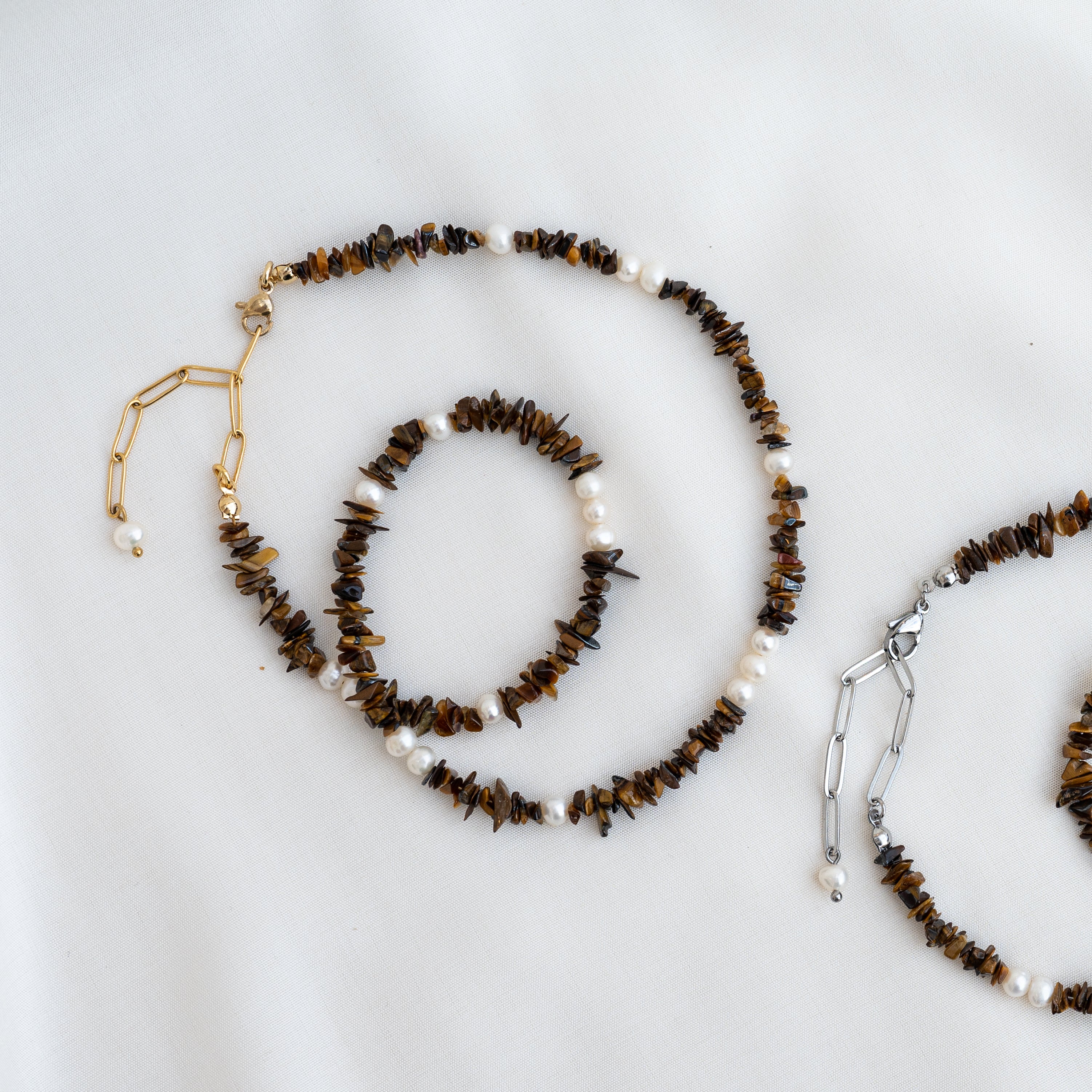 Eine Halskette und ein Armband mit braunen Steinen und weißen Perlen sind auf weißem Stoff angeordnet. Die Halskette hat eine goldene Kette, während das angrenzende Armband und die Kette silberfarben sind.