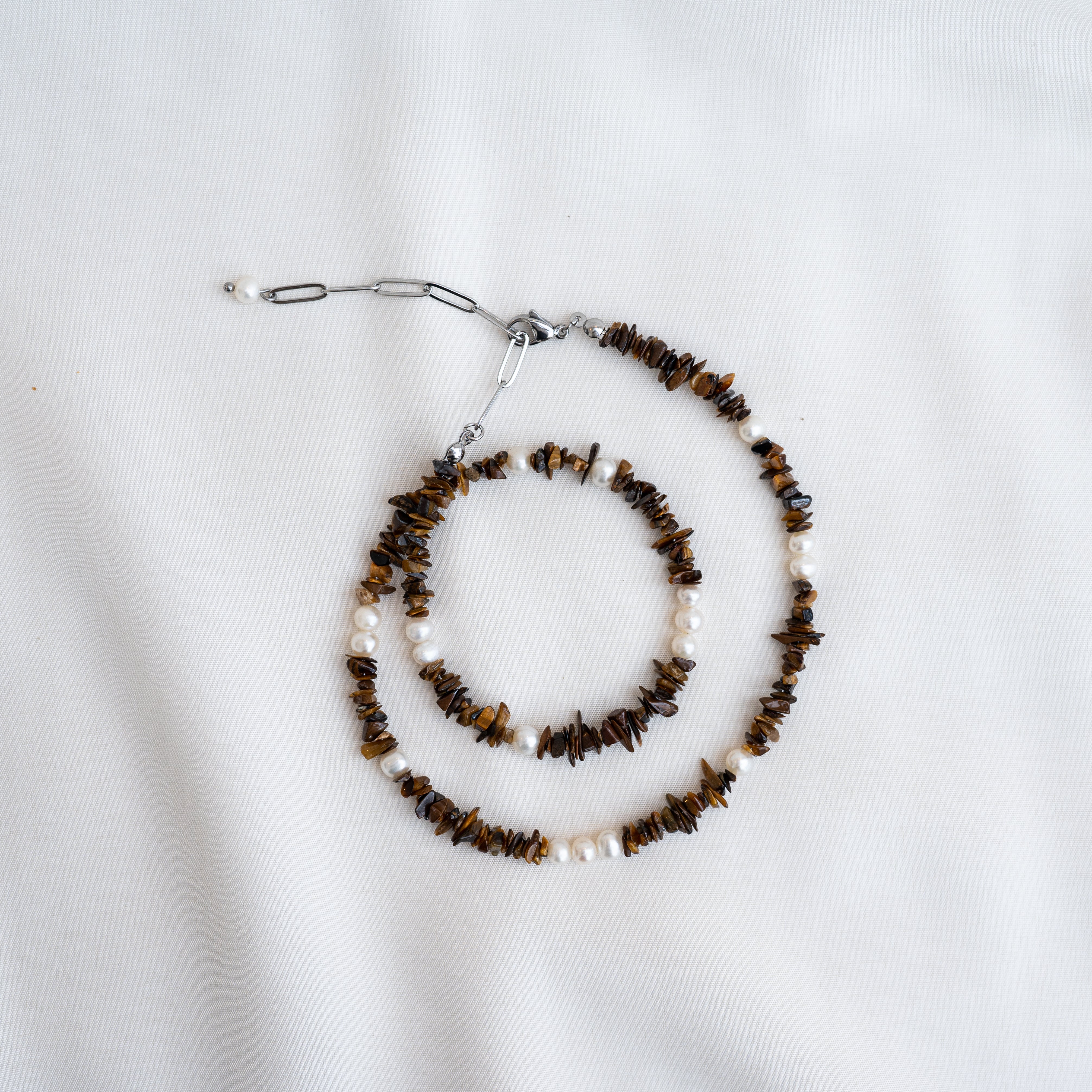 Eine Perlenkette mit abwechselnd braunen Steinsplittern und weißen Perlen, die spiralförmig auf einem weißen Stoffgrund angeordnet sind.
