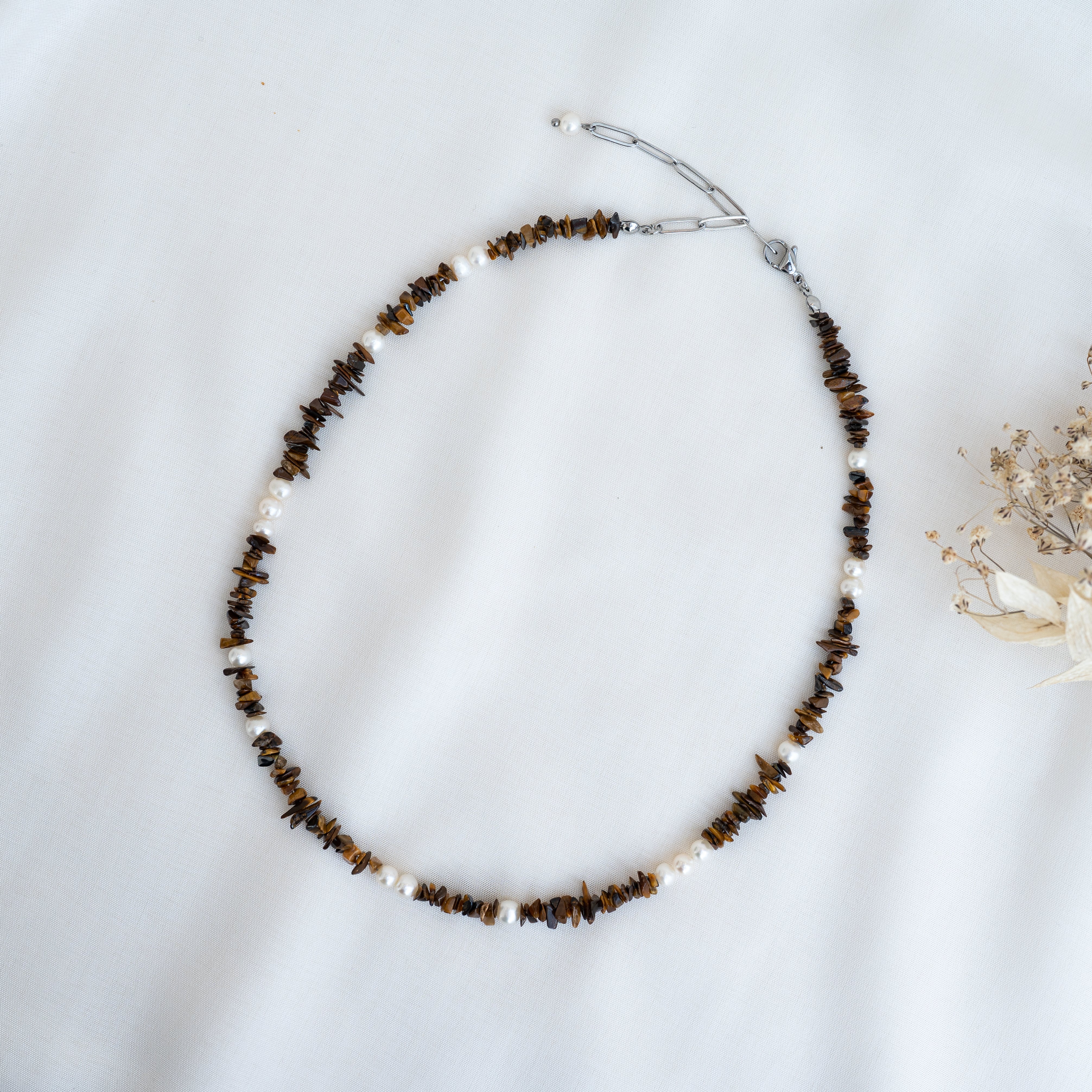 Eine Perlenkette mit kleinen braunen Steinen und weißen Perlen ist kreisförmig auf einer weißen Stofffläche angeordnet, rechts daneben befinden sich Trockenblumen.