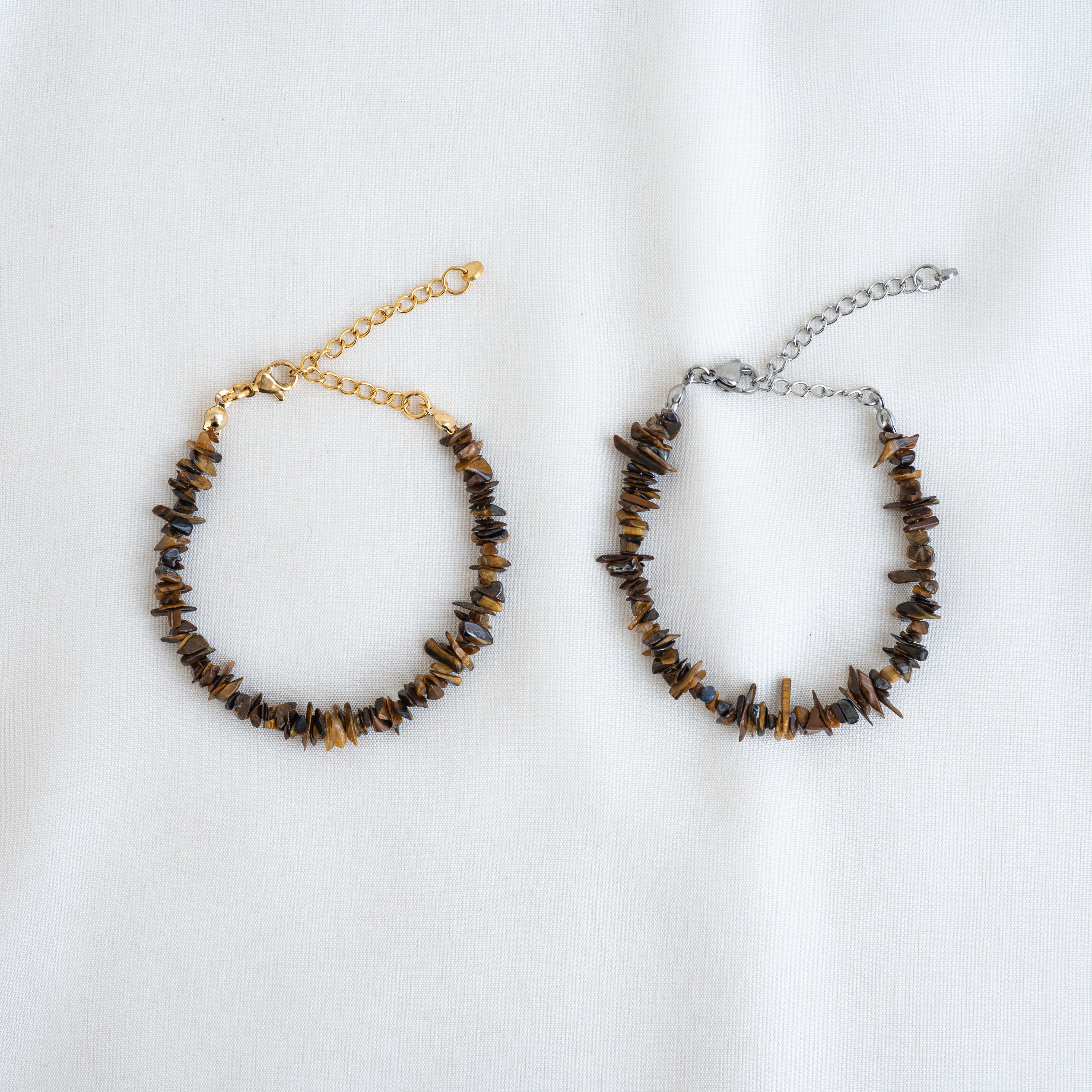 Zwei Armbänder aus unregelmäßigen braunen Steinperlen sind auf einer weißen Fläche ausgestellt. Ein Armband ist mit einer goldenen Kette und einem Verschluss versehen, das andere mit einer silbernen Kette und einem Verschluss.