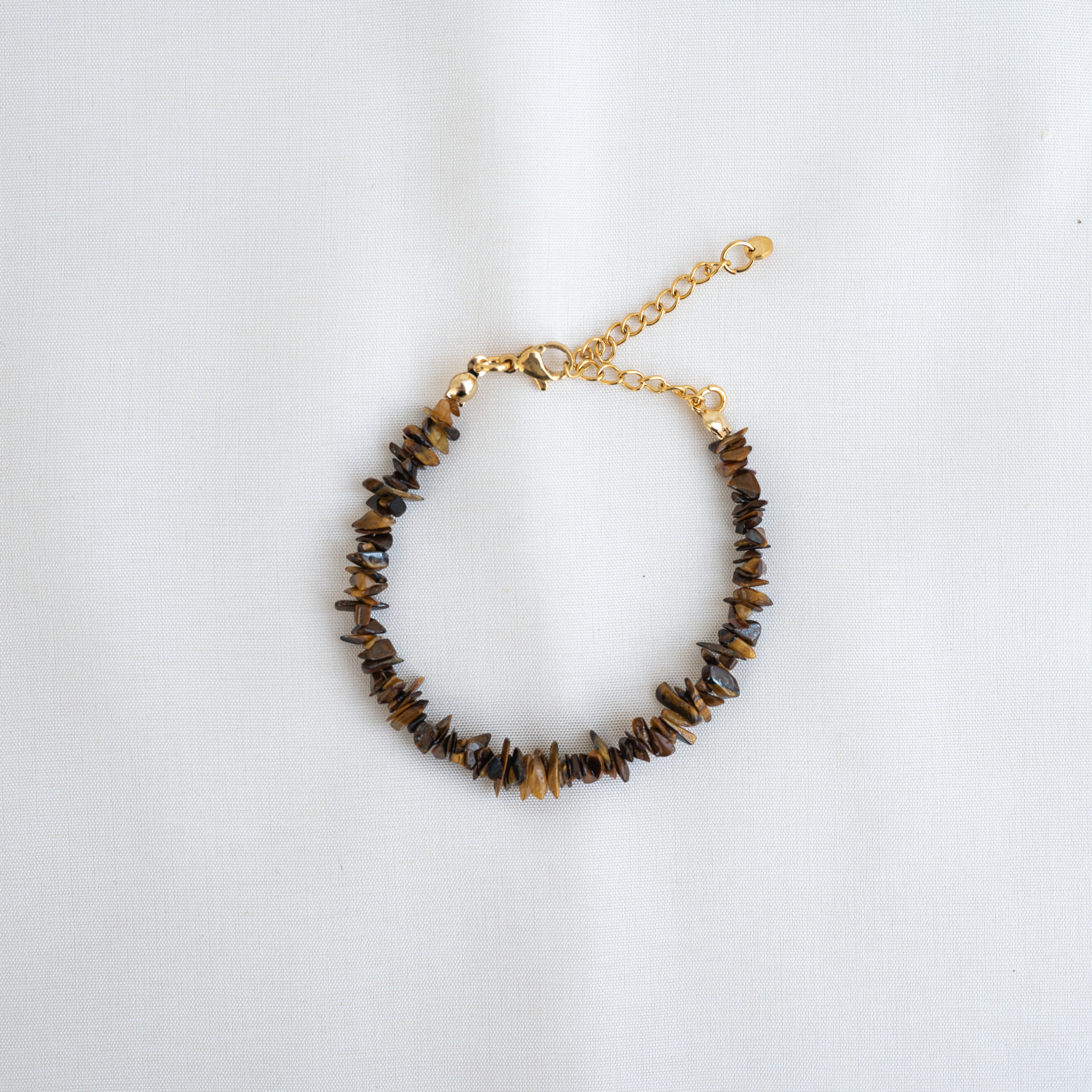 Ein Perlenarmband mit unregelmäßigen braunen Steinsplittern und goldener Kette, mit Karabinerverschluss und verstellbarer Verlängerung, auf weißem Stoffgrund.