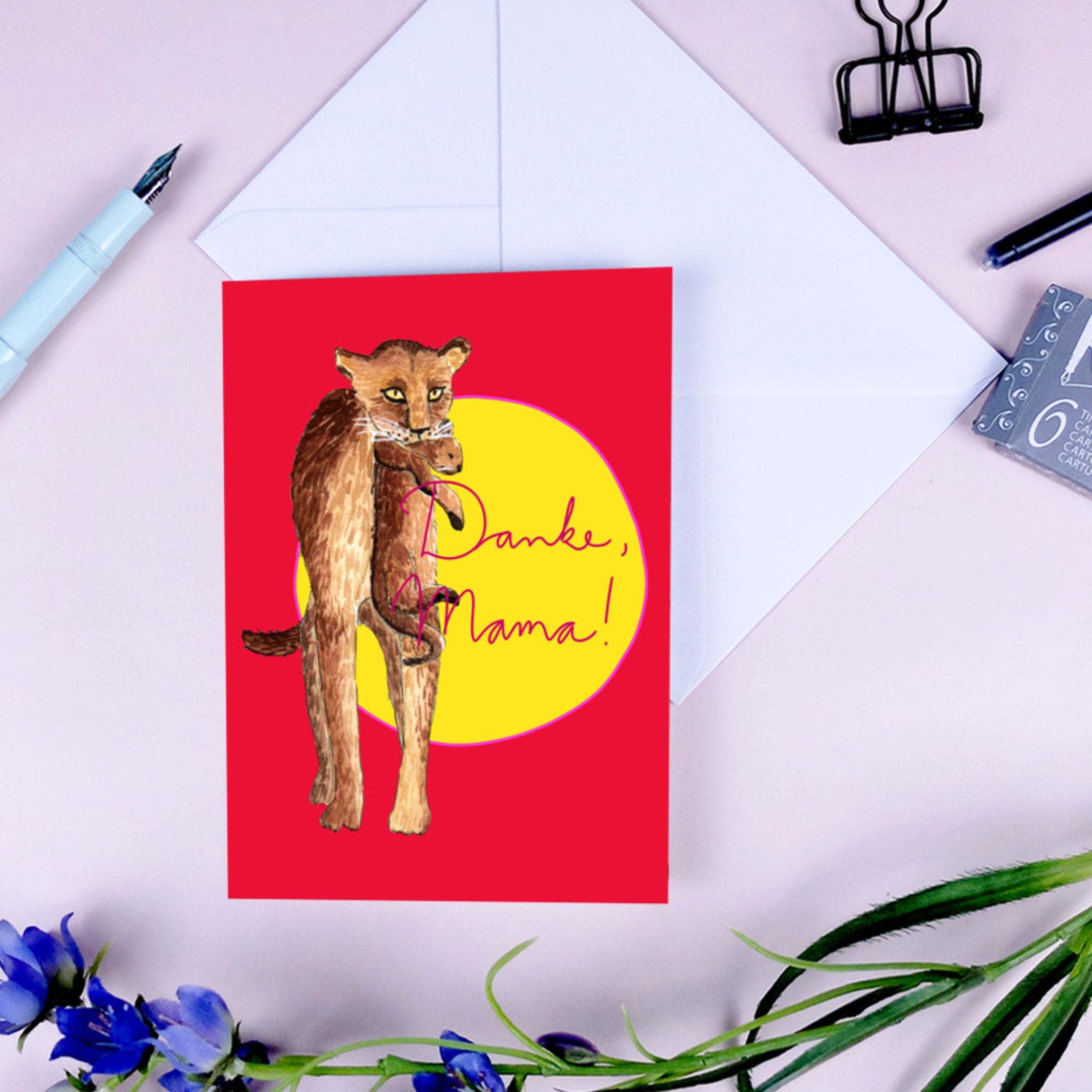 Karte mit einem Löwenjunges und dem Schriftzug „Danke, Mama!“ auf rotem Hintergrund, umgeben von Stift und Blumen.