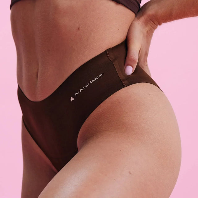 Eine Person in einem braunen Crop-Top und passender hochtaillierter Unterwäsche posiert vor einem rosa Hintergrund. Auf der Unterwäsche steht der Text The Female Company.