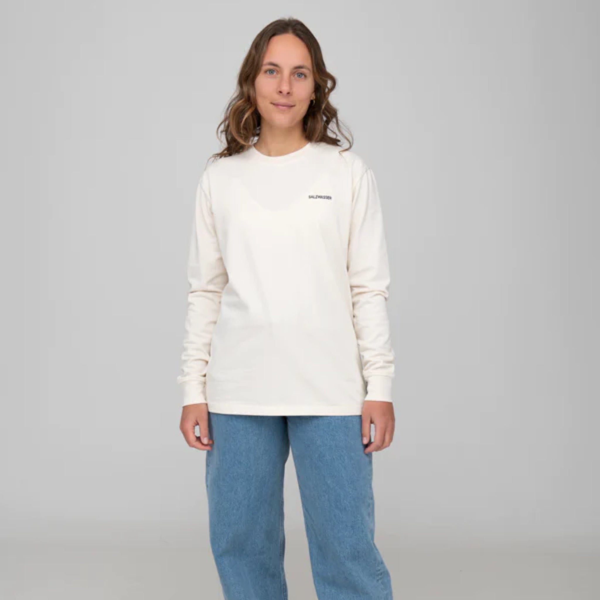 Eine Person trägt ein langärmeliges, cremefarbenes Shirt und blaue Jeans vor grauem Hintergrund.