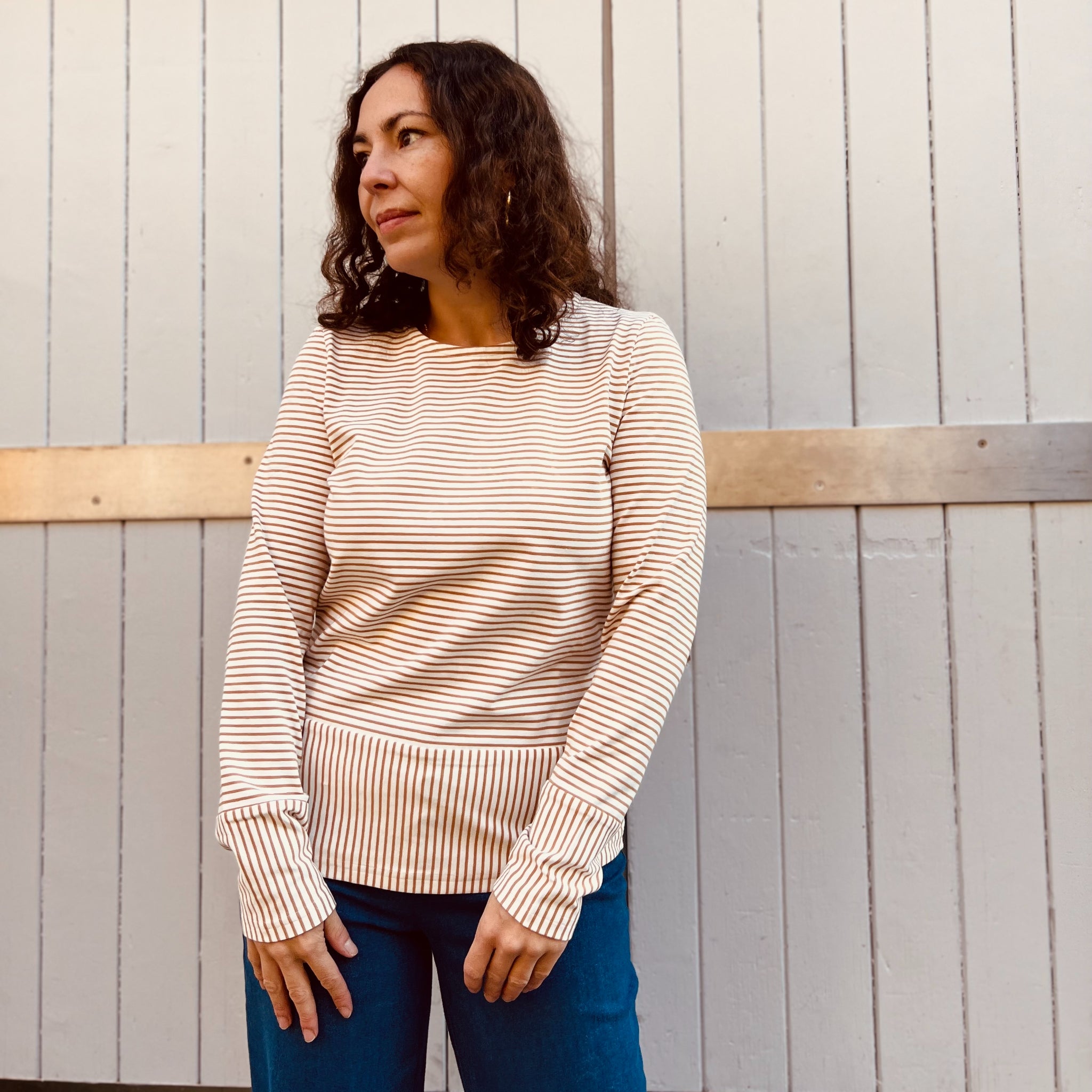 Frau mit lockigem Haar trägt ein gestreiftes Oberteil und blaue Jeans, posiert vor einer vertäfelten Holzwand.