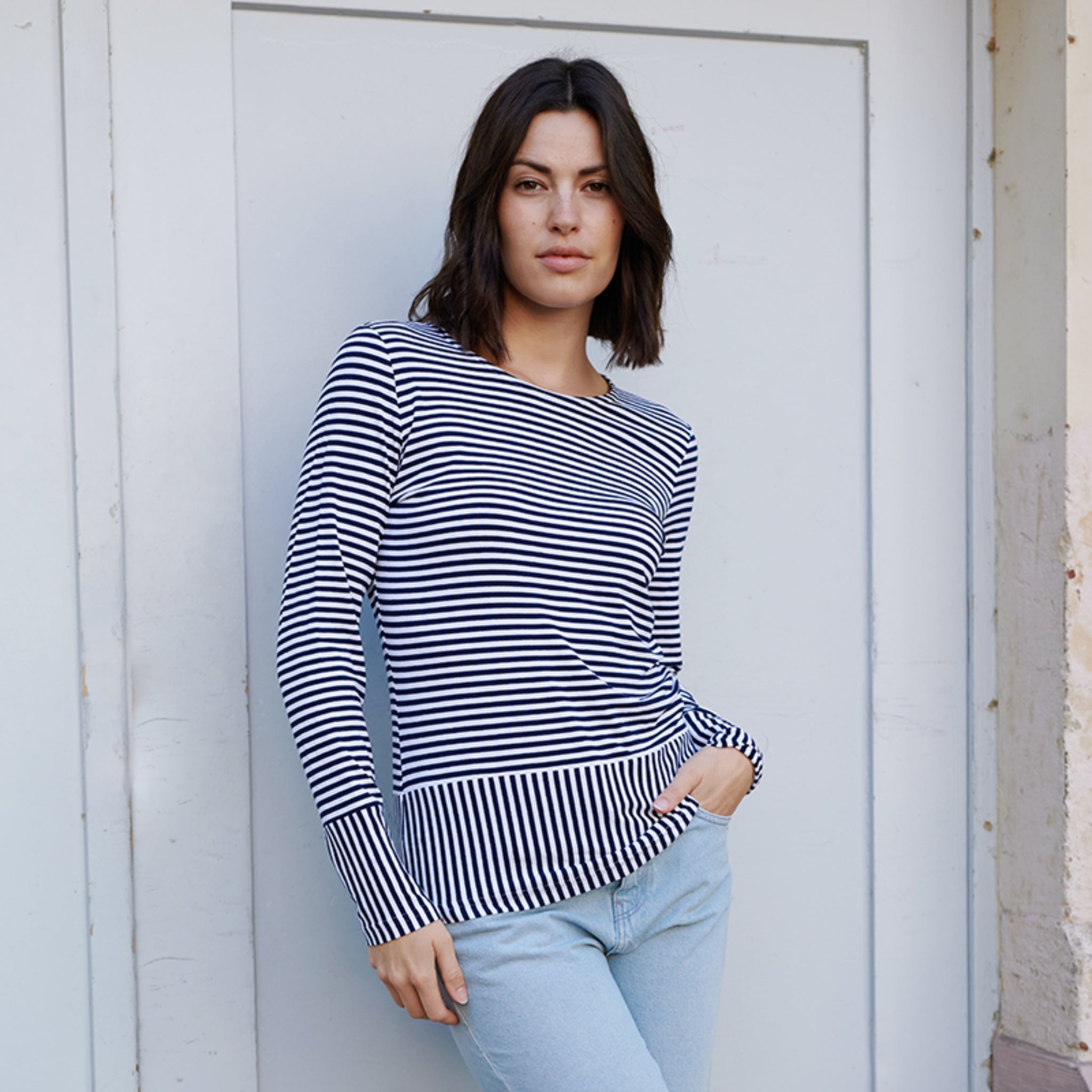 Frau in gestreiftem Pullover lehnt sich an eine helle Wand und trägt eine hellblaue Jeans.