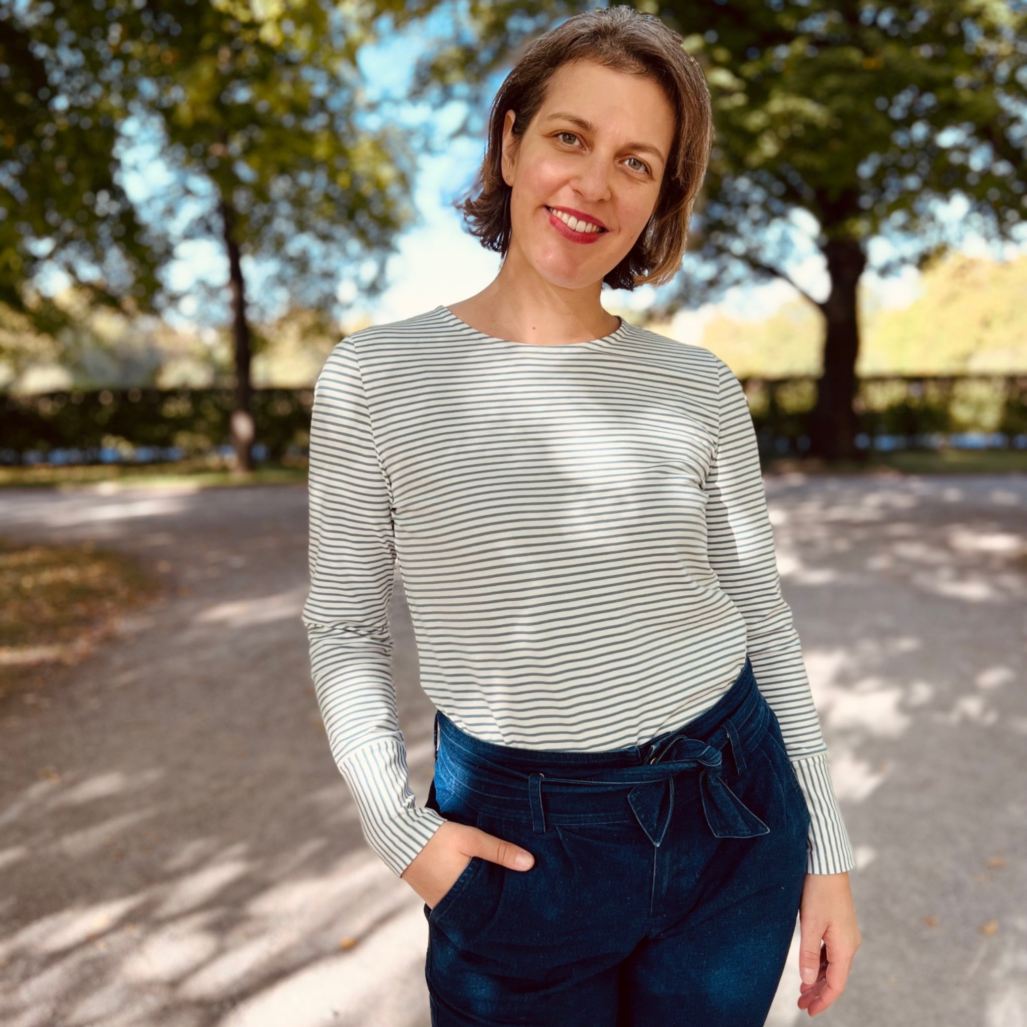 Frau in gestreiftem Oberteil und Jeans steht lächelnd auf einem sonnigen Weg im Park.