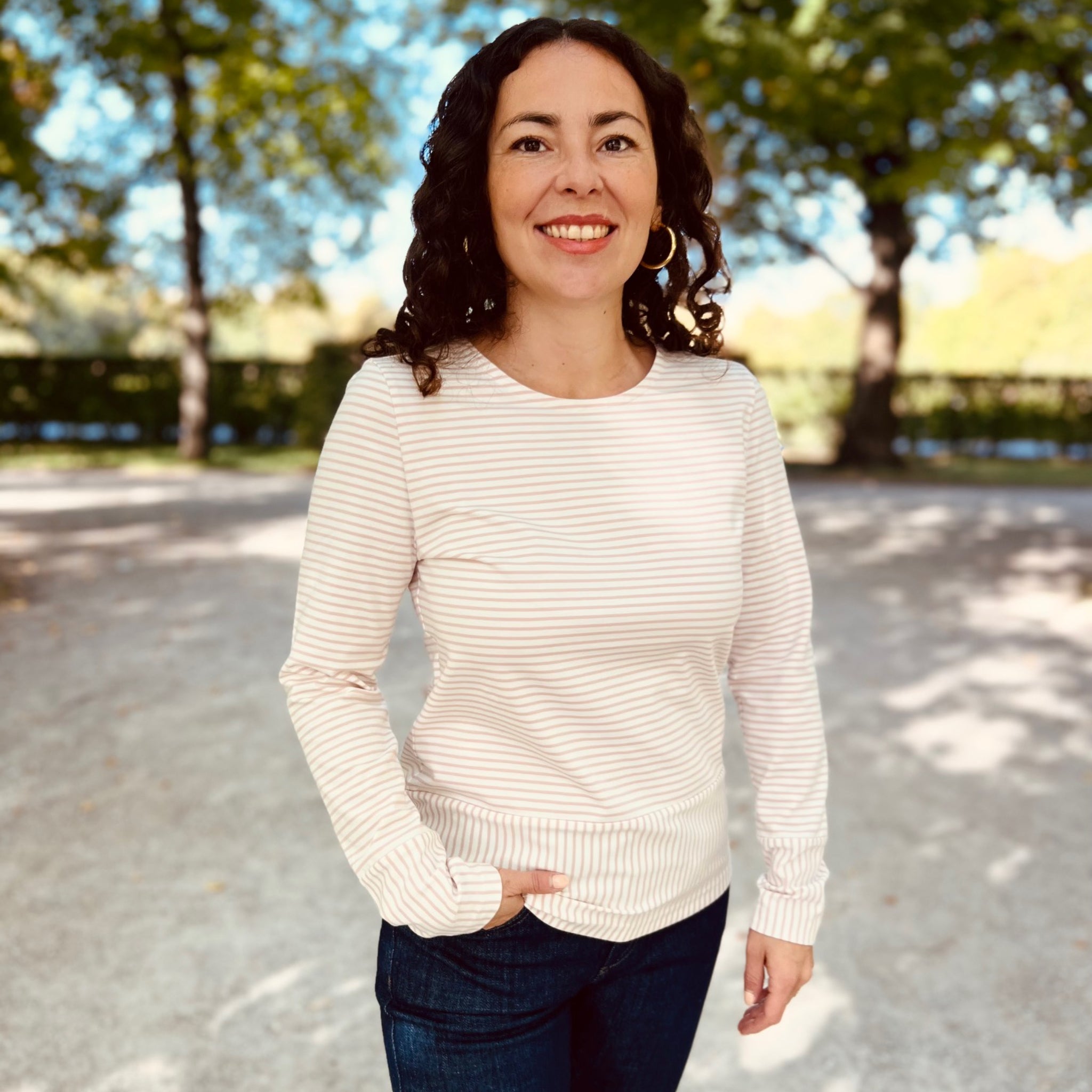 Eine Frau mit lockigem Haar lächelt im Freien, trägt ein gestreiftes Oberteil und Jeans, und steht vor Bäumen.