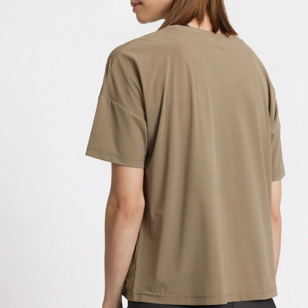 Person in beigem T-Shirt von hinten, mit schulterlangem braunem Haar, vor weißem Hintergrund.