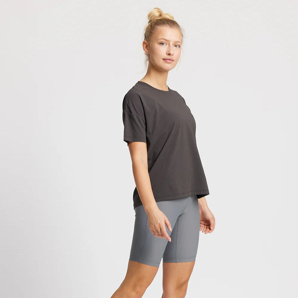 Eine Person trägt ein schwarzes T-Shirt und graue Radlerhose, steht vor einem weißen Hintergrund.