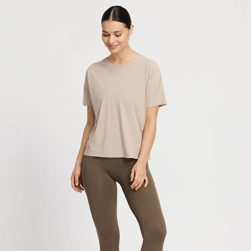 Eine lächelnde Frau trägt ein beigefarbenes T-Shirt und braune Leggings vor einem weißen Hintergrund.
