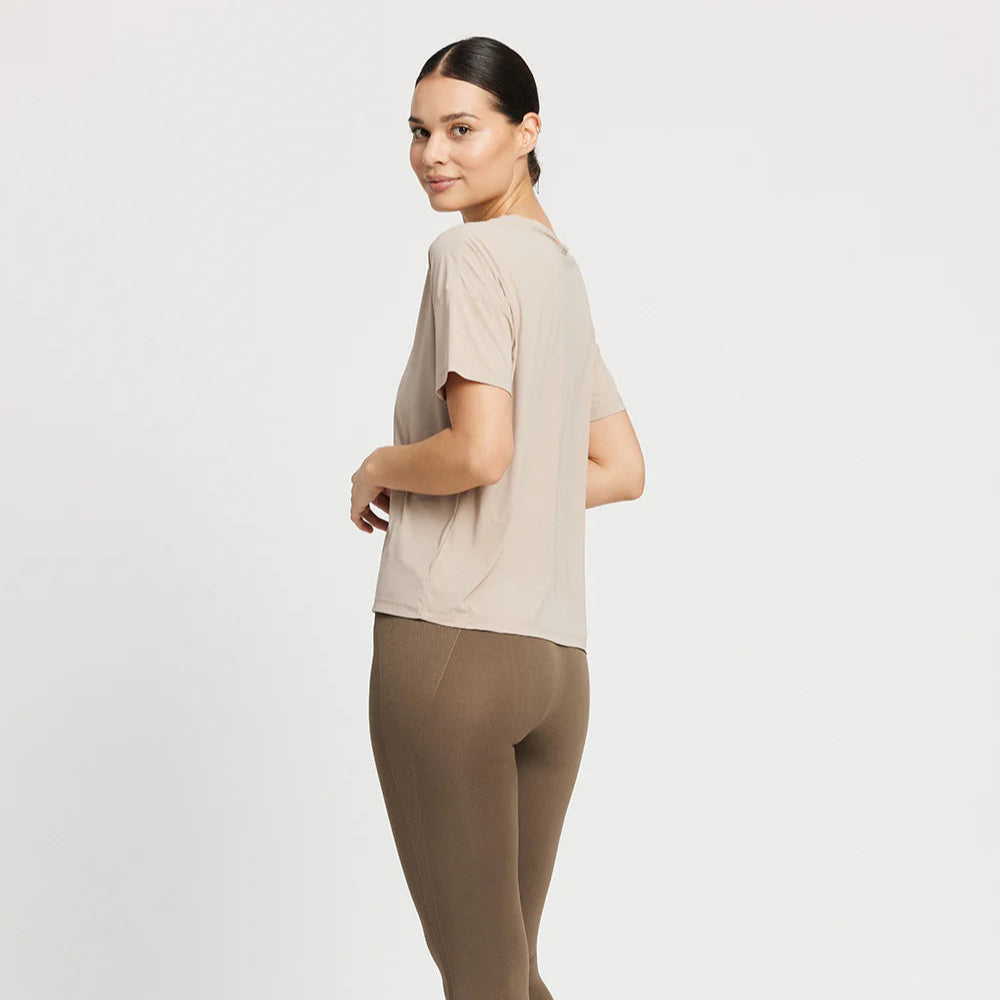 Frau in beigefarbenem Shirt und brauner Leggings, steht mit leicht gedrehter Körperhaltung und lächelt in die Kamera.