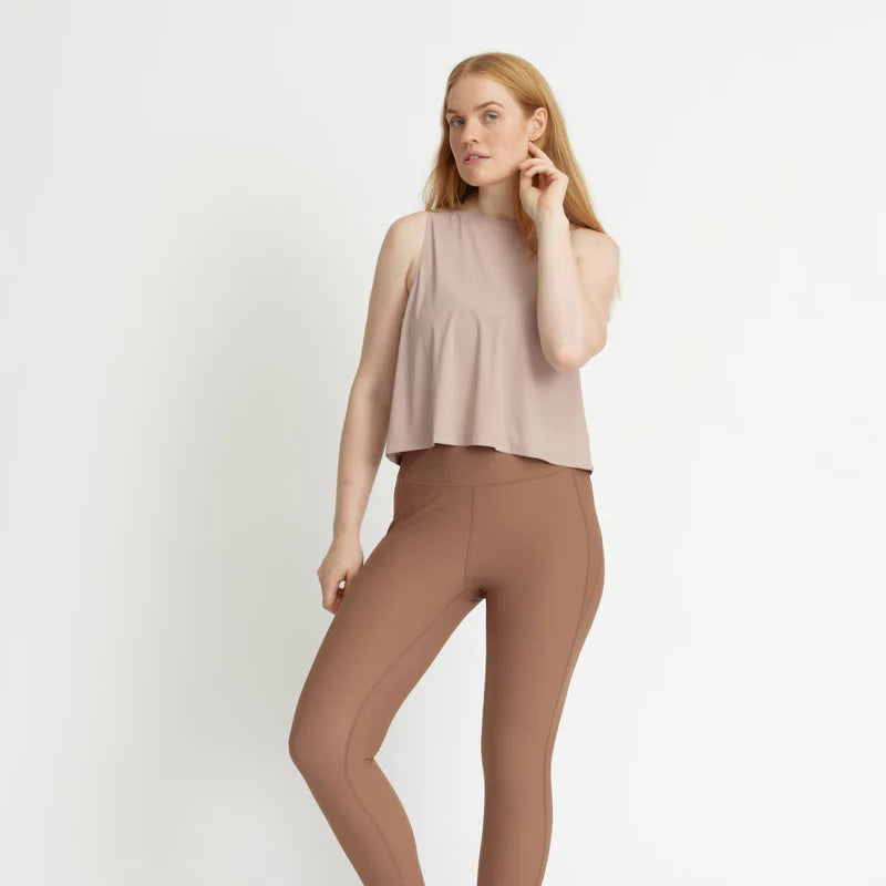 Eine Frau steht vor einem schlichten weißen Hintergrund, trägt ein ärmelloses, lockeres beiges Oberteil und hochtaillierte, eng anliegende braune Leggings, während sie mit einer Hand ihr Haar berührt und die andere auf ihrer Hüfte ruht.