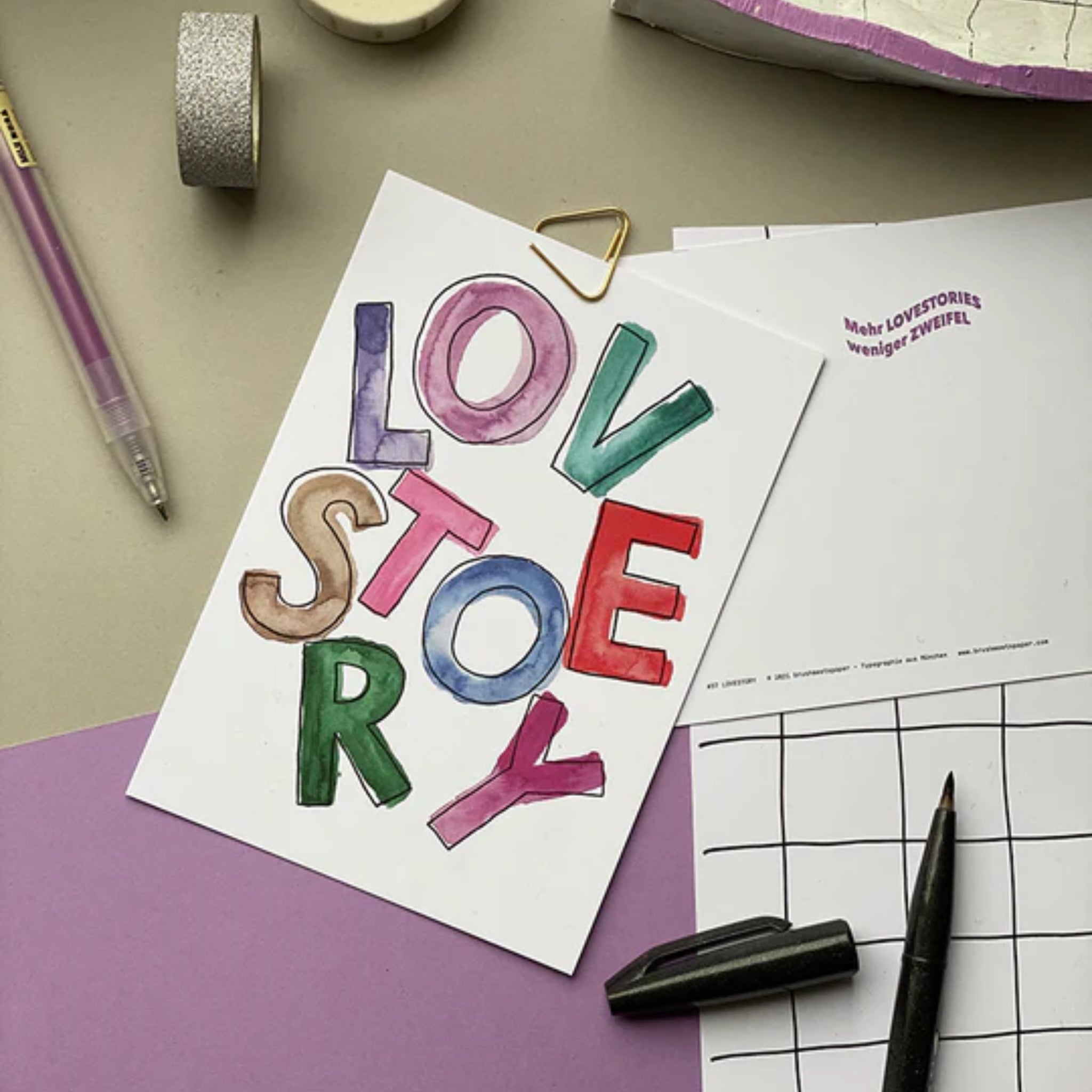 Eine handgemalte Karte mit dem Schriftzug "LOVE STORY" in bunten Buchstaben, daneben ein Stift und lila Papier.