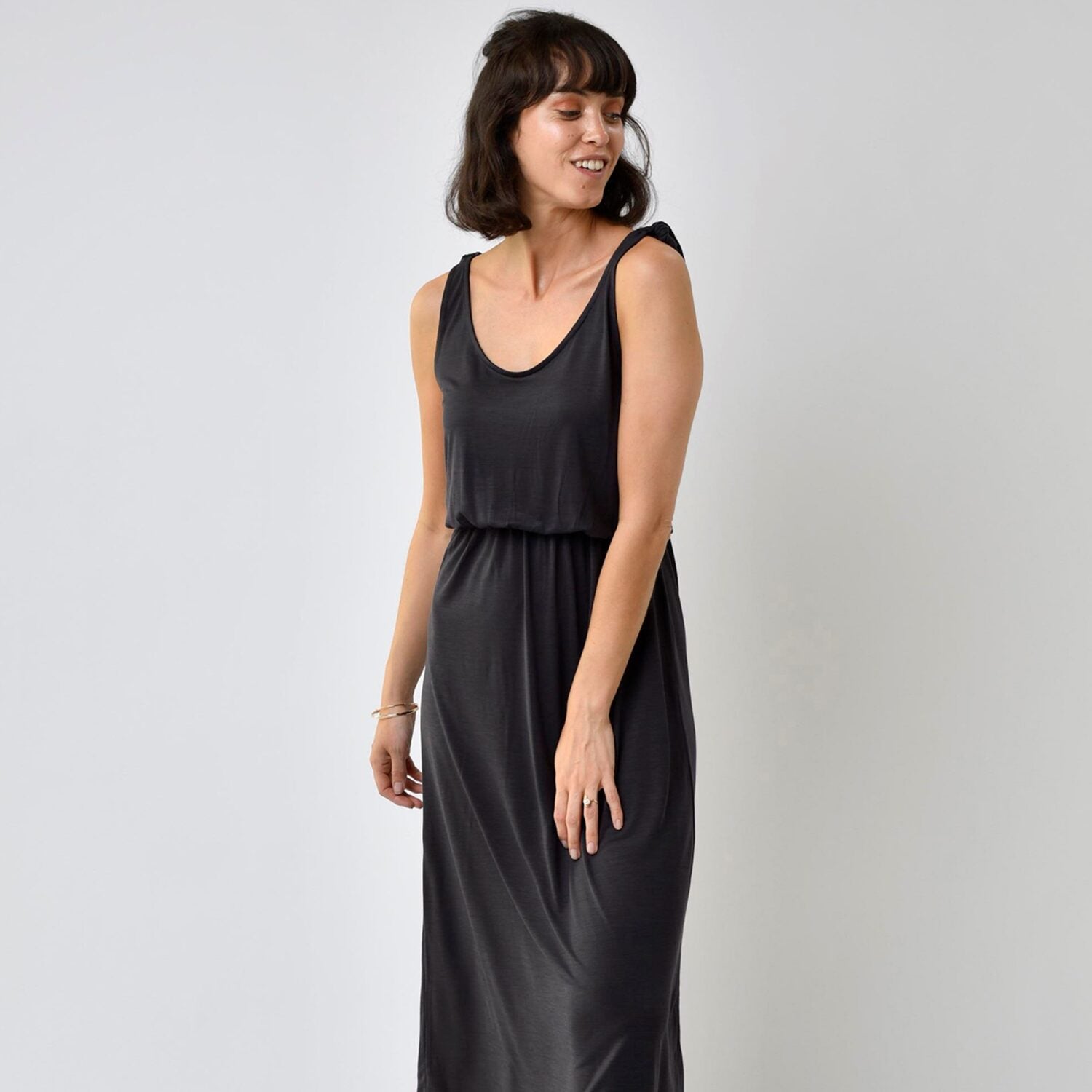 Frau in einem ärmellosen, schwarzen Kleid steht vor einem hellgrauen Hintergrund und lächelt zur Seite.