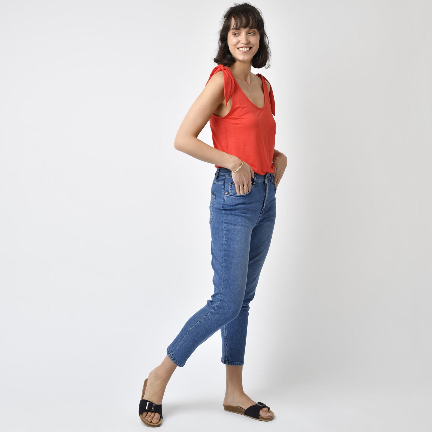 Frau in rotem Tanktop und blauen Jeans posiert lächelnd, mit den Händen in den Hosentaschen, auf weißem Hintergrund.