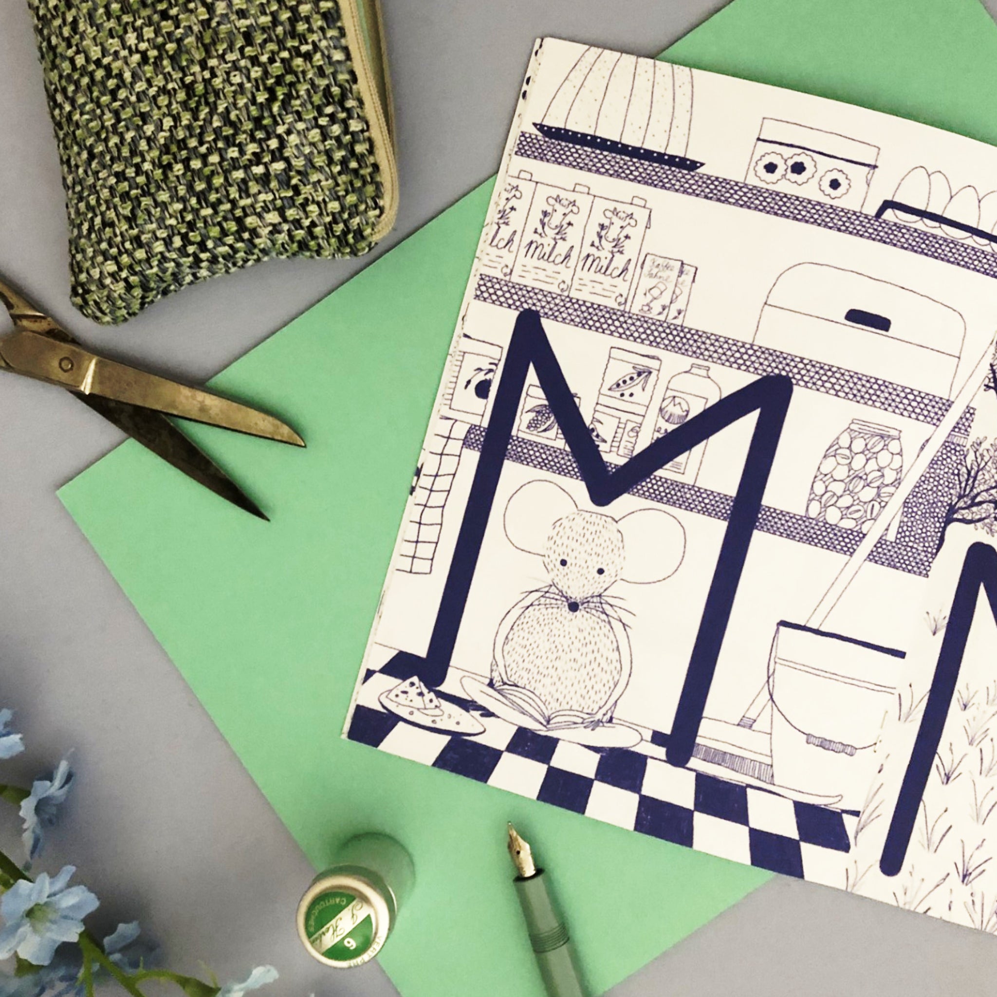 Ein Buch mit einer illustrierten Maus liegt auf grünem Papier neben einer Schere, einem Stoffbeutel und einem Stift.
