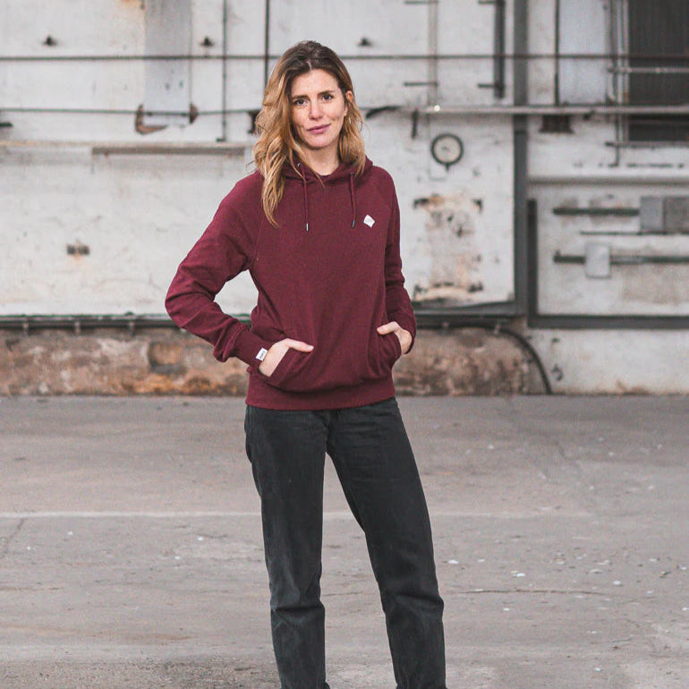 Person in rotem Hoodie und schwarzer Hose steht vor einer industriellen Wand und blickt in die Kamera.