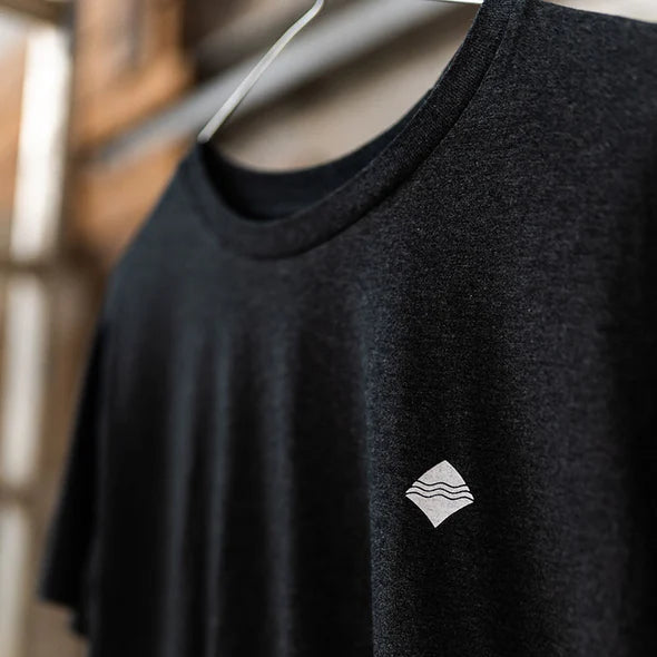 Schwarzes T-Shirt auf einem Kleiderbügel mit kleinem, weißen Logo auf der Brust; im Hintergrund unscharf.