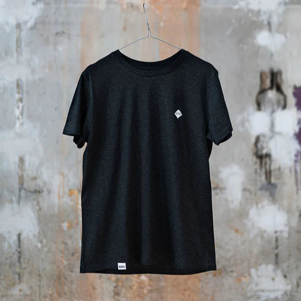Schwarzes T-Shirt auf einem Kleiderbügel vor einer betonartigen, farbig unregelmäßigen Wand aufgehängt.