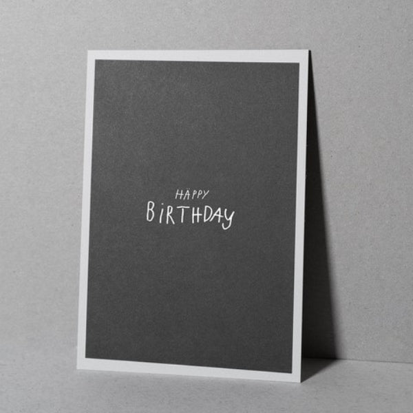 Eine minimalistische Geburtstagskarte mit schwarzer Vorderseite und weißem Rand, auf der die Worte HAPPY BIRTHDAY in einfachen weißen handgeschriebenen Buchstaben aufrecht vor einem grauen Hintergrund stehen.