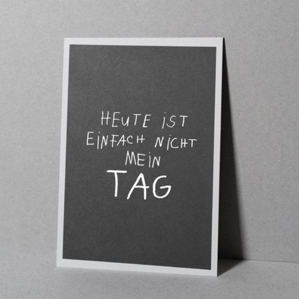 Eine schwarze Karte mit weißem handgeschriebenem Text auf Deutsch, der Heute ist einfach nicht mein Tag lautet, steht vor einem grauen Hintergrund.