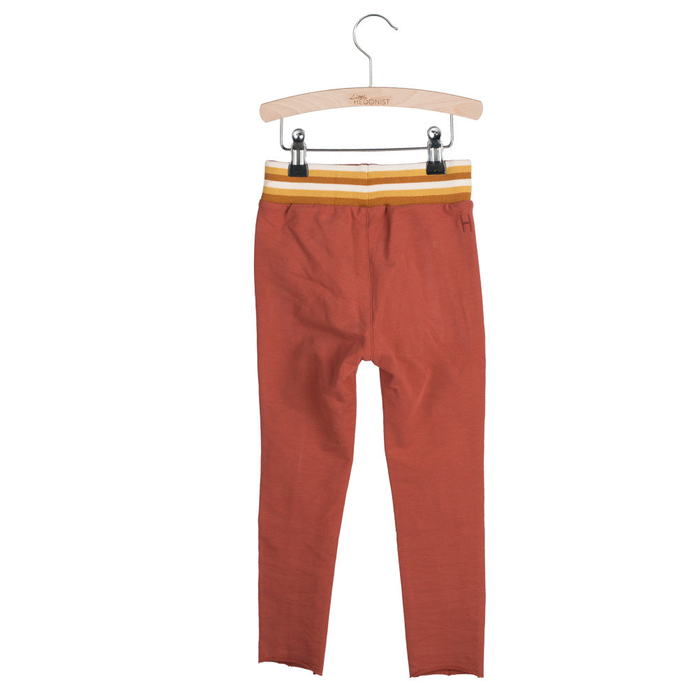 Rostfarbene Kinderleggings mit gelb-weiß gestreiftem Gummibund, ausgestellt auf einem Holzbügel vor weißem Hintergrund.