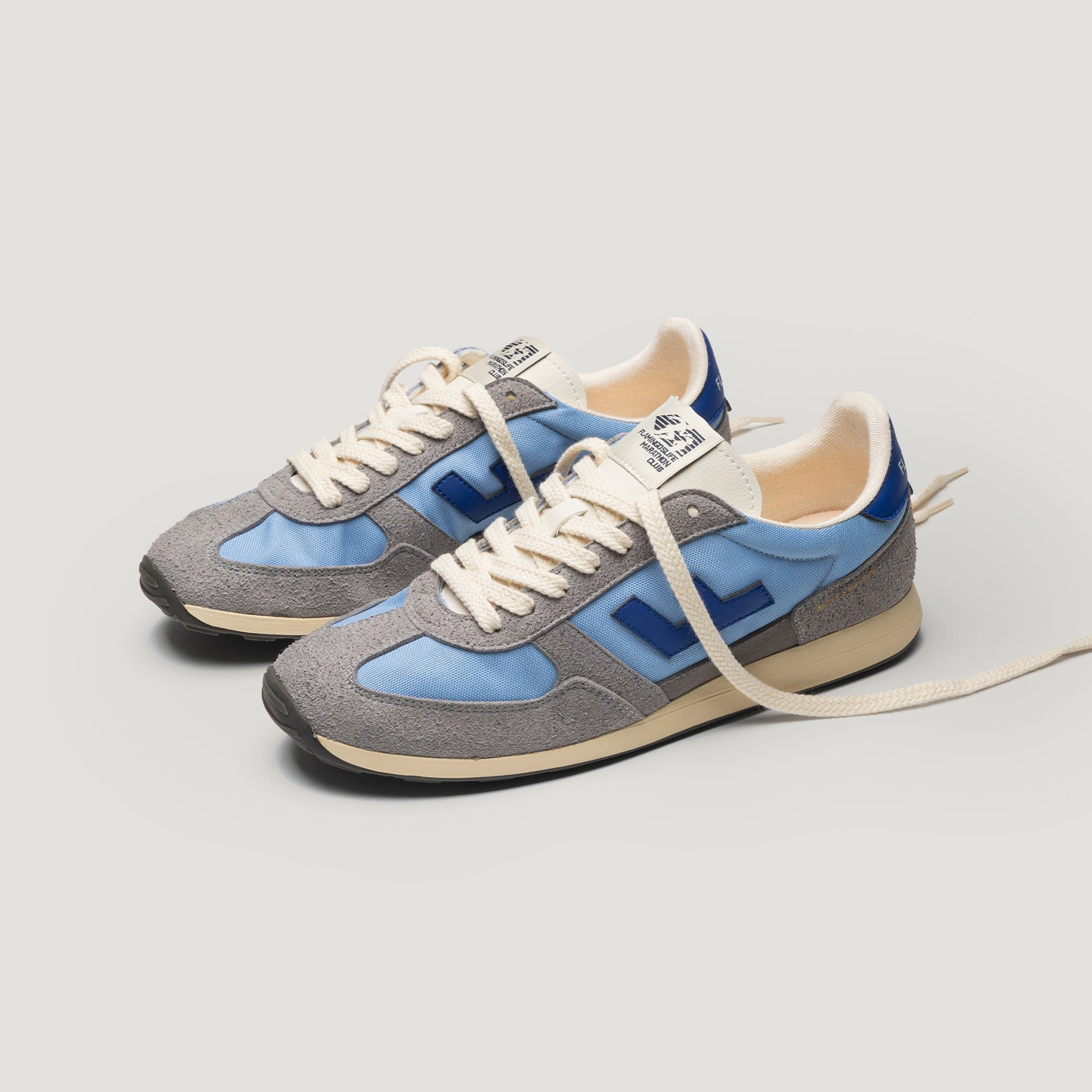 Ein Paar stilvolle Turnschuhe mit grauen, blauen und marineblauen Einsätzen, cremefarbenen Schnürsenkeln und cremefarbenen Zwischensohlen vor einem schlichten hellen Hintergrund. Ein Schnürsenkel ist gelöst und nach außen verlängert.