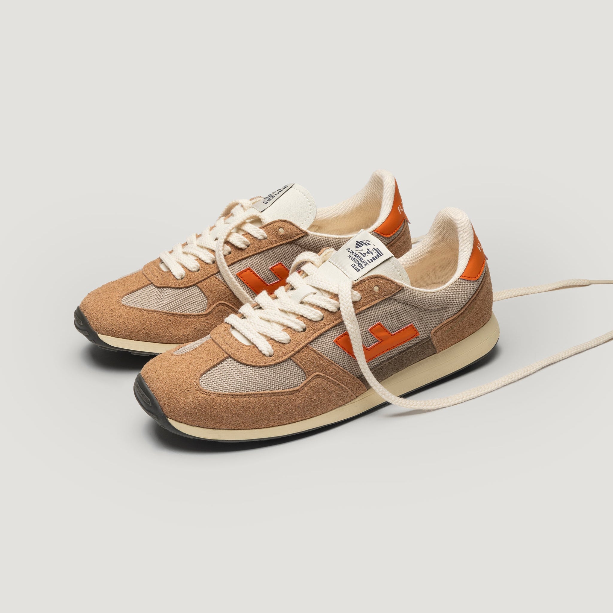 Ein Paar beigefarbene und braune Turnschuhe mit orangefarbenen Akzenten, weißen Schnürsenkeln und schwarzen Sohlen, die nebeneinander auf einem schlichten hellen Hintergrund stehen. Ein Schnürsenkel ist locker nach außen gezogen.