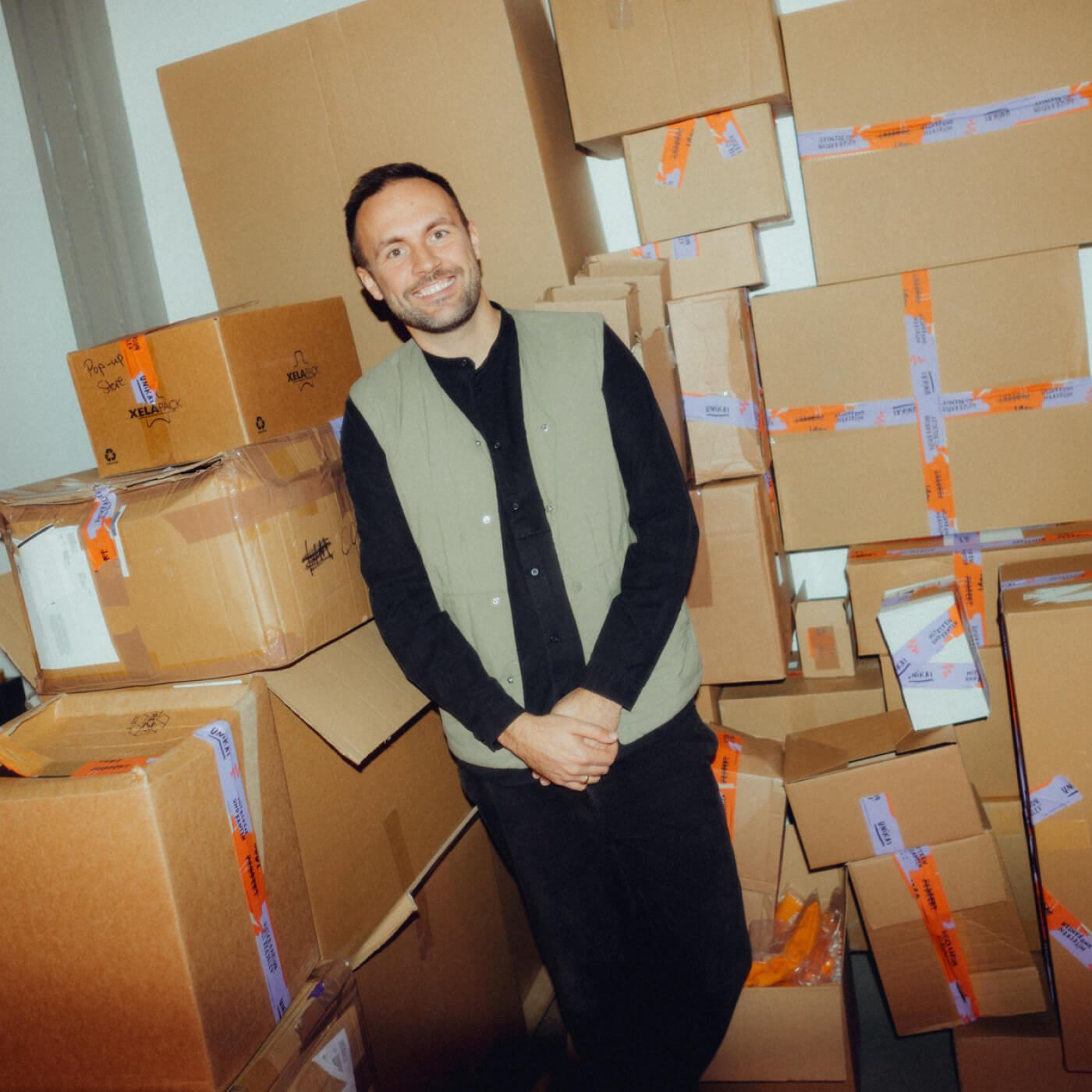 Ein Mann steht lächelnd vor vielen gestapelten Kartons.
