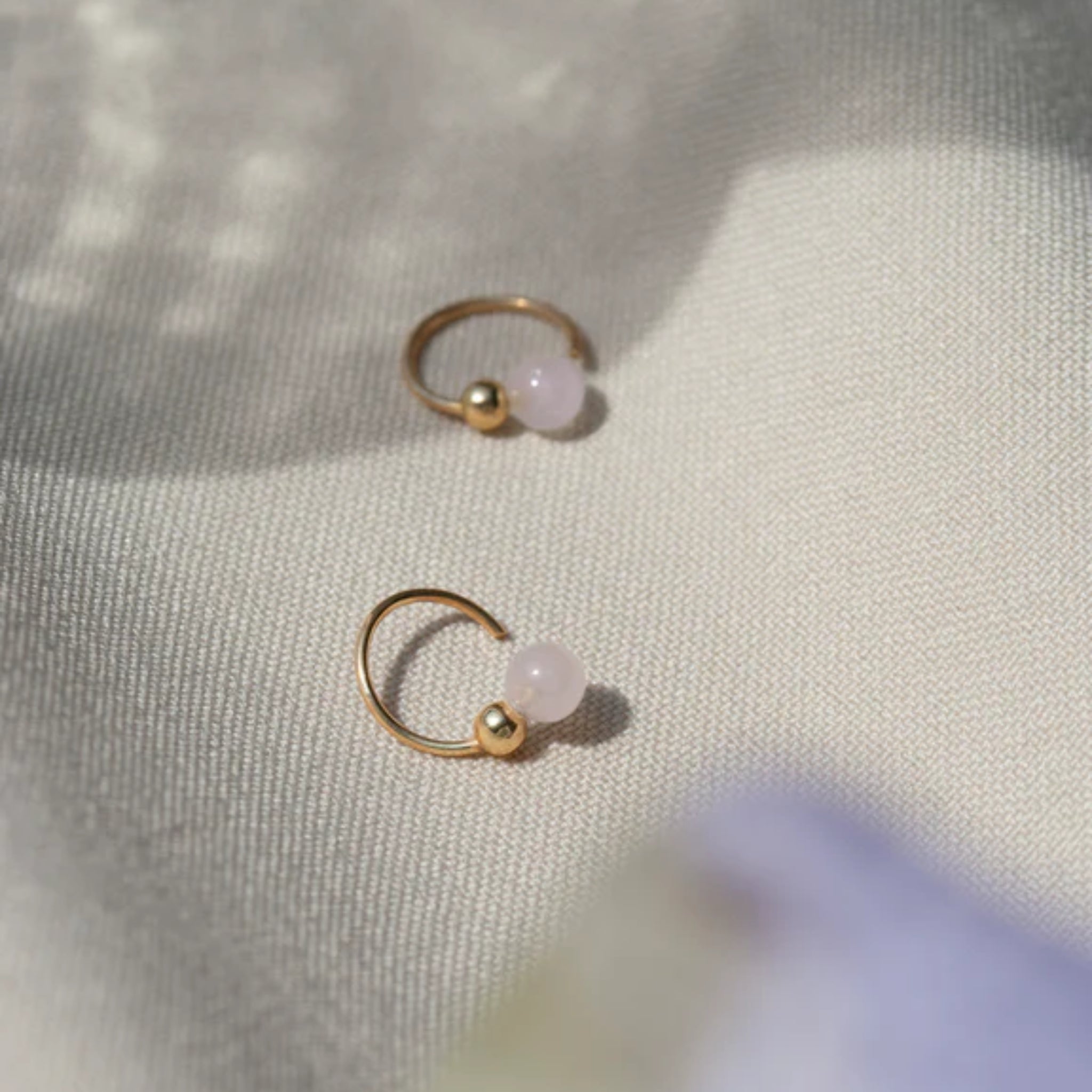 Zwei goldene Ohrringe mit kleinen rosa Perlen liegen auf einem cremefarbenen Stoff im Sonnenlicht.