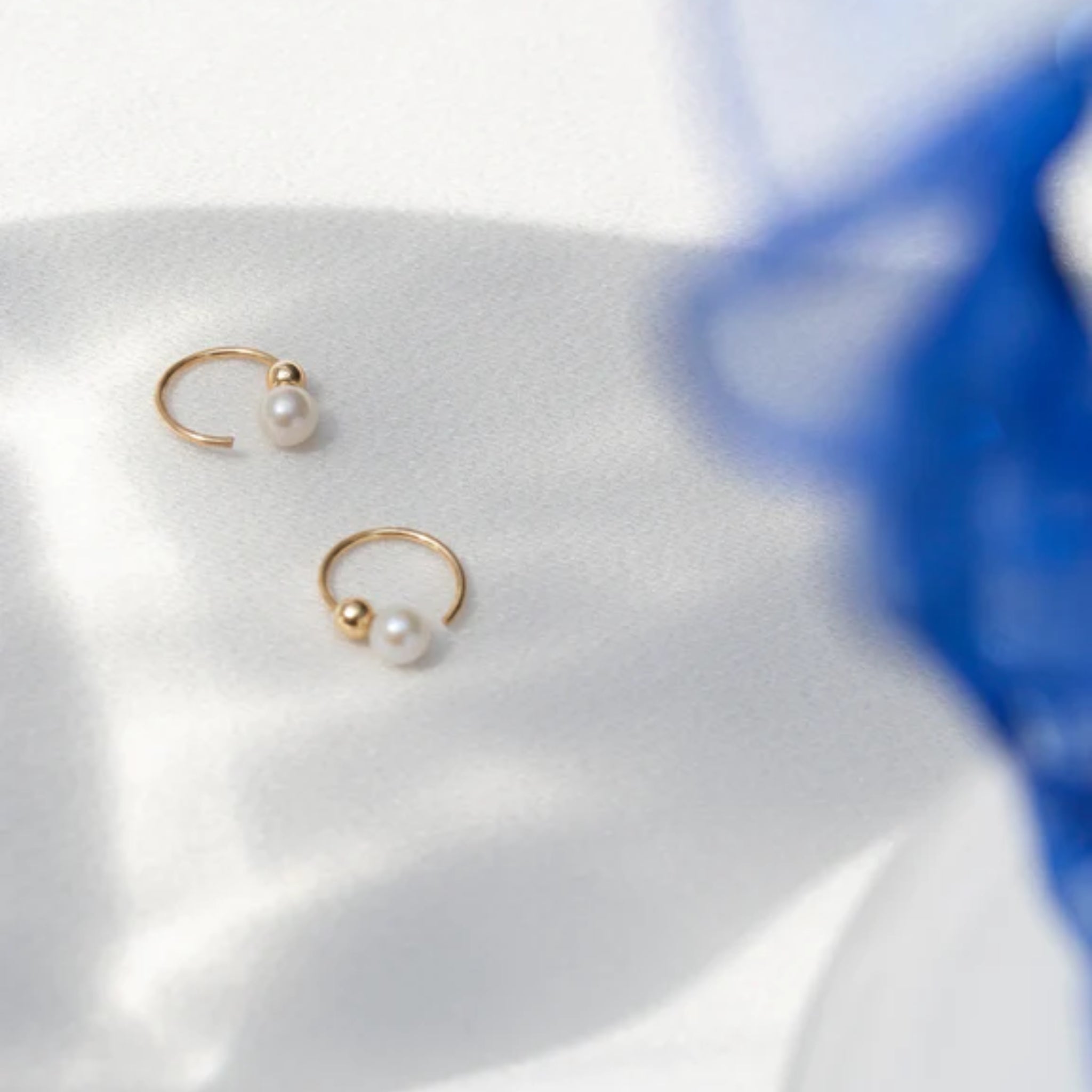 Zwei goldene Ohrringe mit Perlen liegen auf weißem Stoff, daneben ein unscharfer blauer Gegenstand.