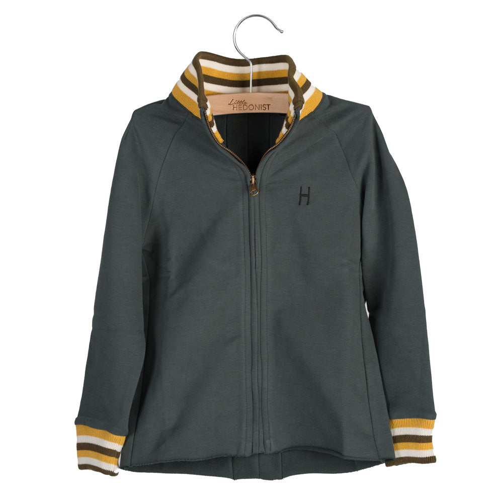 Dunkelgrüne Jacke mit Reißverschluss und gelb-weiß gestreiften Manschetten und Kragen, die auf einem Bügel hängt. Die Jacke hat ein kleines H-Logo auf der linken Brust.