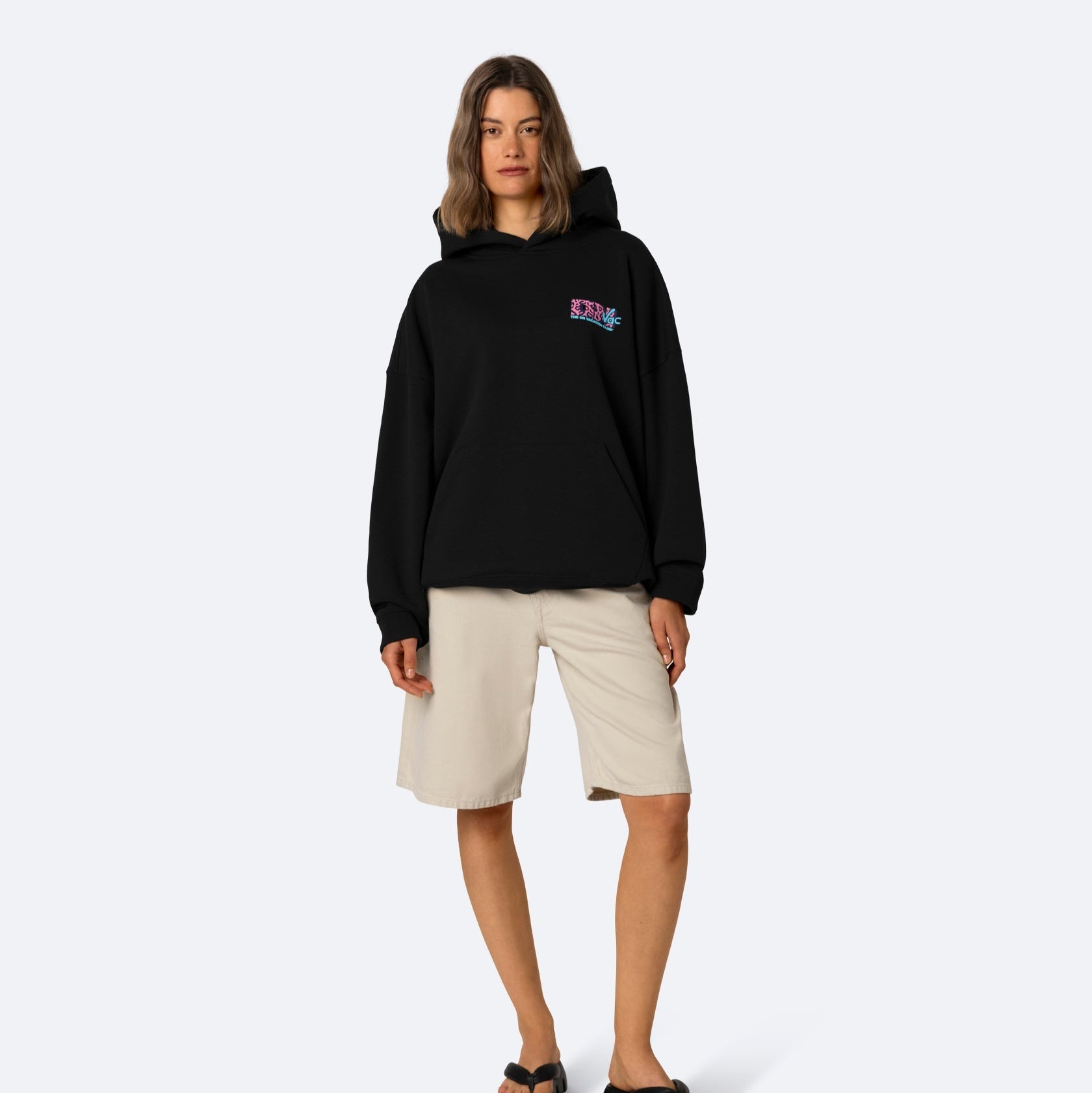 Eine Frau steht vor einem schlichten Hintergrund und trägt einen schwarzen Kapuzenpullover mit einer kleinen Grafik auf der Brust, beigefarbene knielange Shorts und schwarze Sandalen. Sie hat schulterlanges Haar und einen neutralen Gesichtsausdruck.