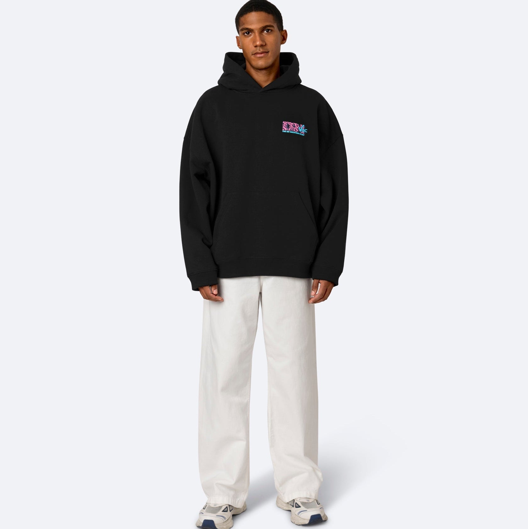 Eine Person steht mit dem Gesicht nach vorne und trägt einen schwarzen Kapuzenpullover mit einer kleinen bunten Grafik auf der Brust, eine weiße Hose mit weitem Bein und weiße Turnschuhe vor einem schlichten hellen Hintergrund.