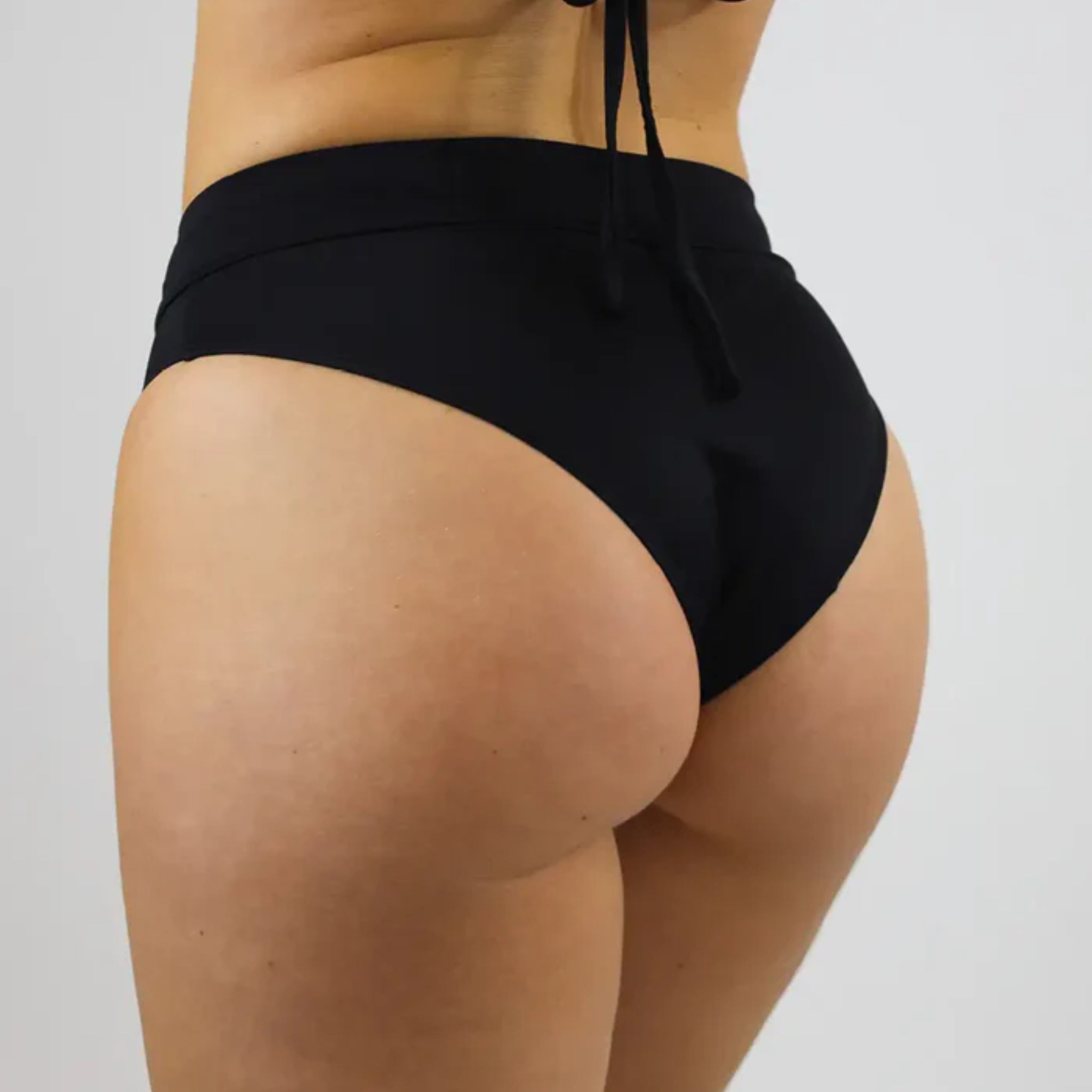 Eine Person trägt schwarze Bikinislips, Rücken zur Kamera, auf grauem Hintergrund.