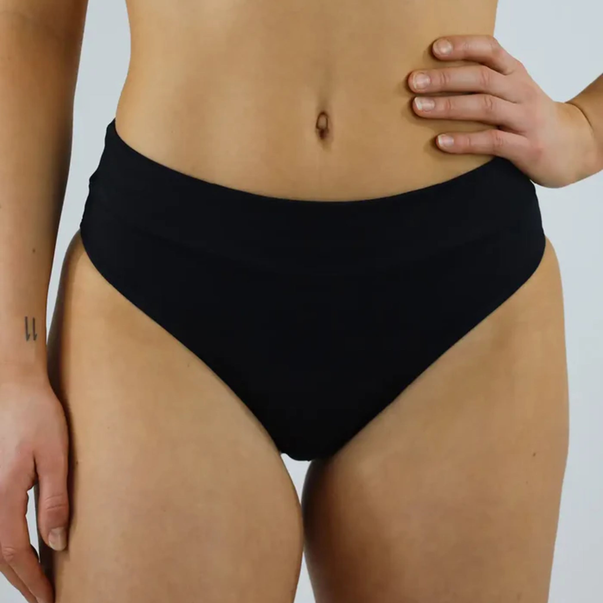 Person trägt einen schwarzen, hochgeschnittenen Badeunterteil; eine Hand auf der Hüfte, eine Tätowierung am Arm.