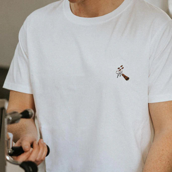 Eine Person, die ein schlichtes weißes T-Shirt mit einem kleinen gestickten Motiv einer Person trägt, die einen Besen hält, steht an einer Kaffeemaschine und bereitet einen Espresso zu.