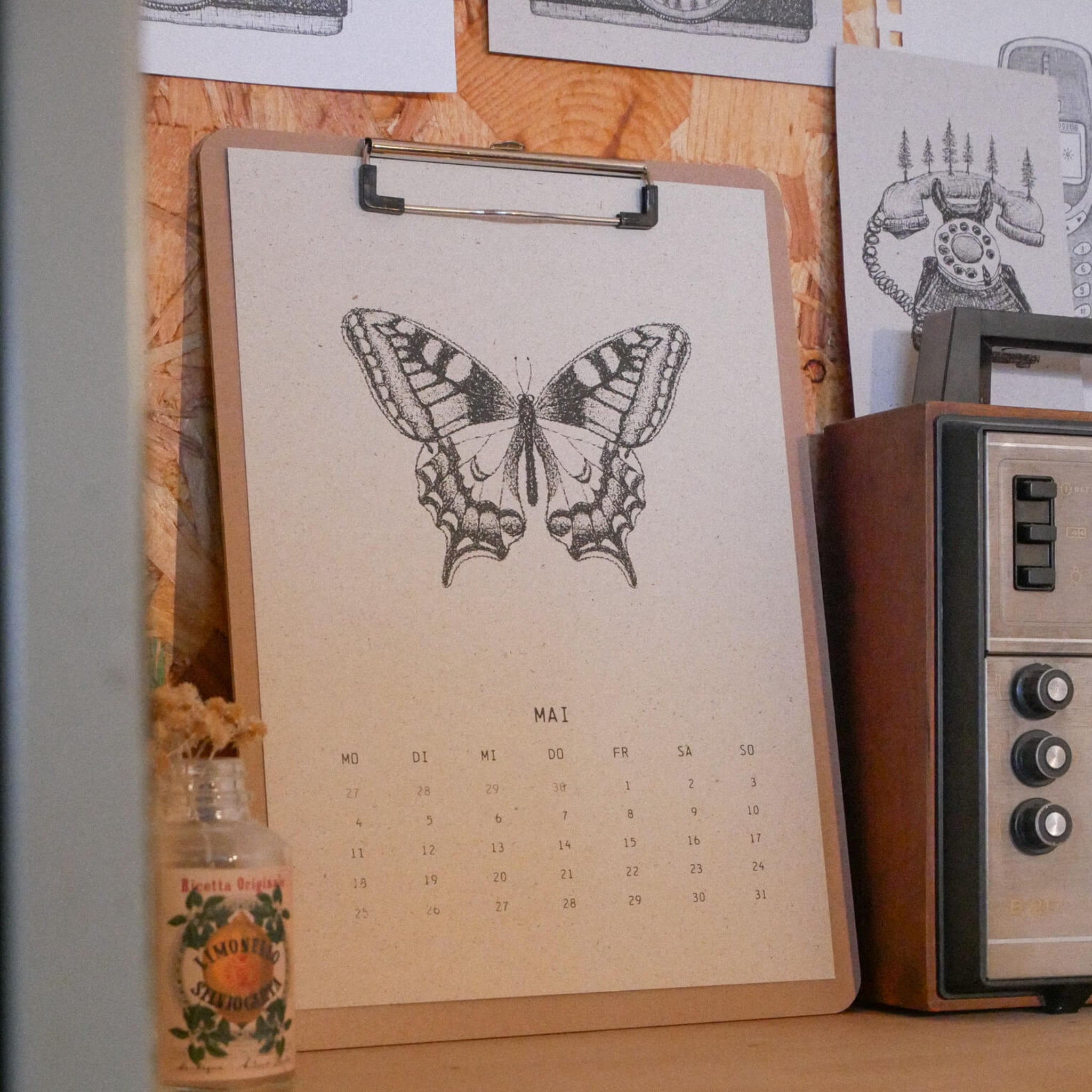 Ein Klemmbrett mit einem Kalenderblatt für den Monat Mai, das eine detaillierte Schmetterlingsillustration zeigt, liegt auf einer hölzernen Oberfläche neben einem alten Radio und einer kleinen Glasflasche mit getrockneten Blumen. Im Hintergrund hängt eine Zeichnung an der Wand.