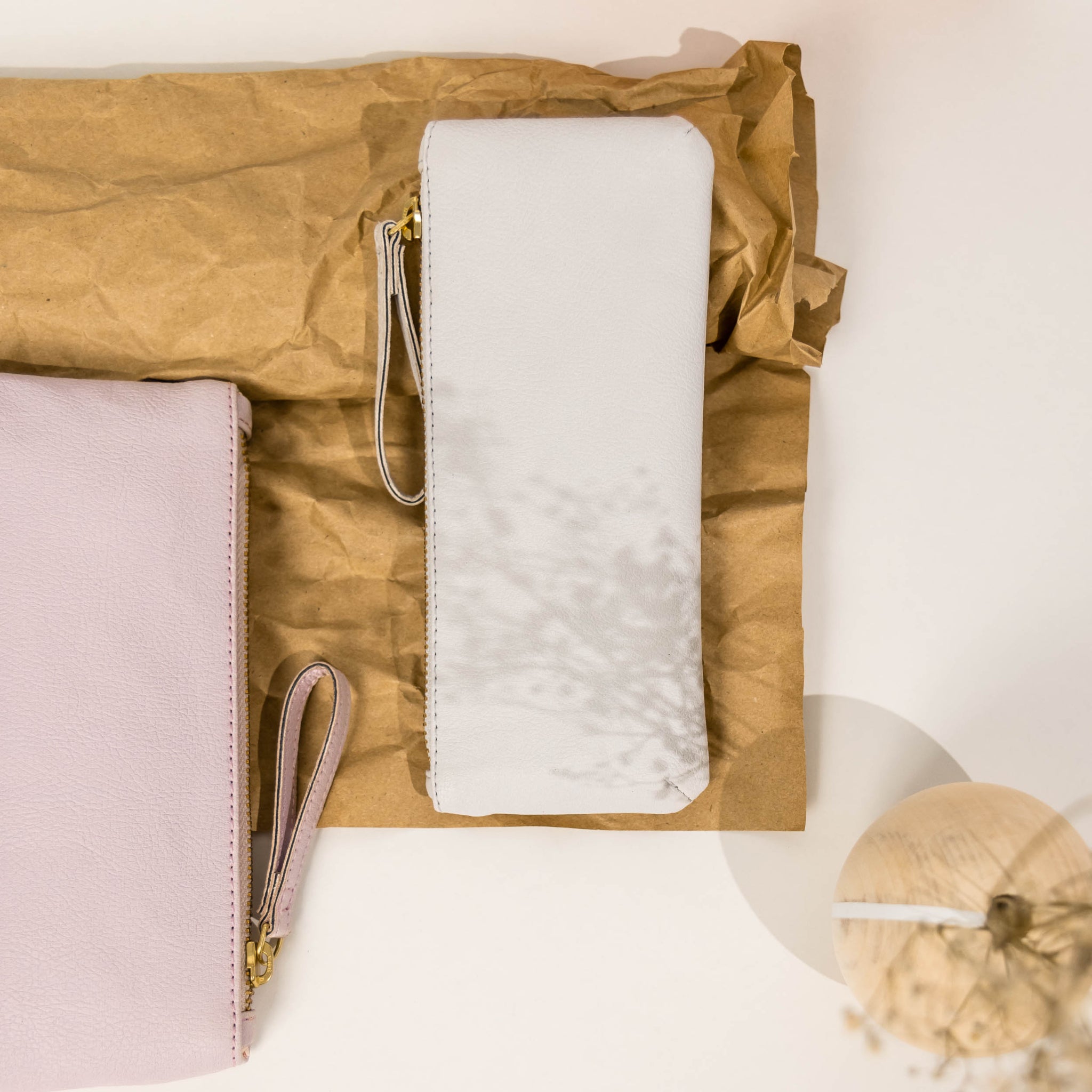 Zwei elegante Ledertaschen, eine beige und eine rosa, auf braunem Papier mit einem dekorativen Zweig im Vordergrund.