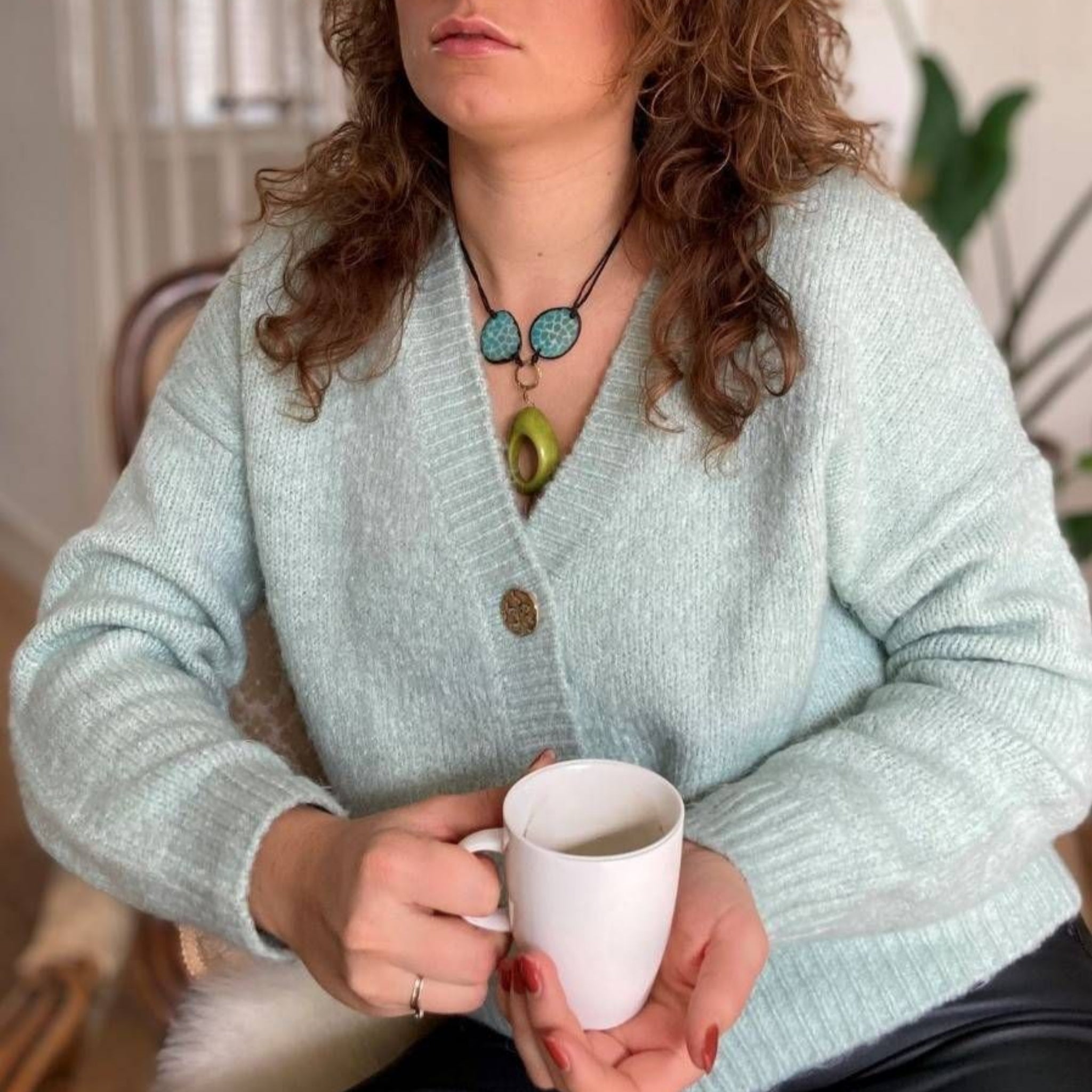 Eine Person mit lockigem Haar, die eine hellblaue Strickjacke trägt, hält eine weiße Tasse. Sie trägt eine türkisfarbene Halskette und einen Avocado-Anhänger. Im Hintergrund sind unscharfe Zimmerpflanzen und Möbel zu sehen.